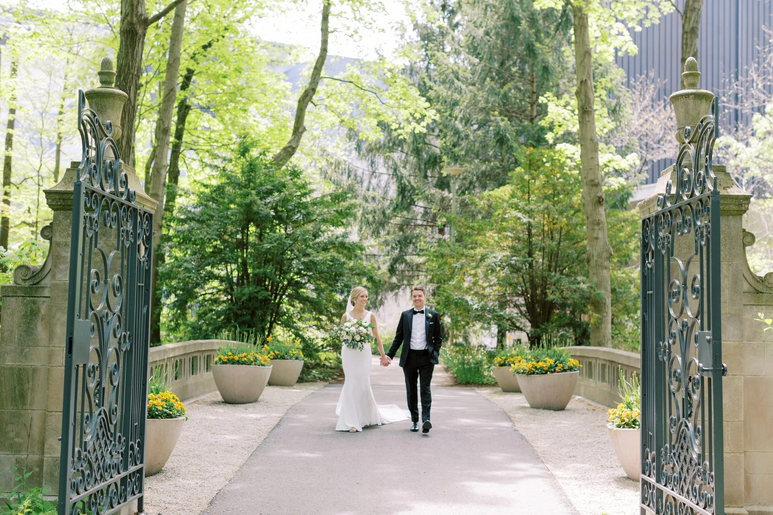 A bride and groom holding hands and walking through a large open gate, with trees and greenery in the background.