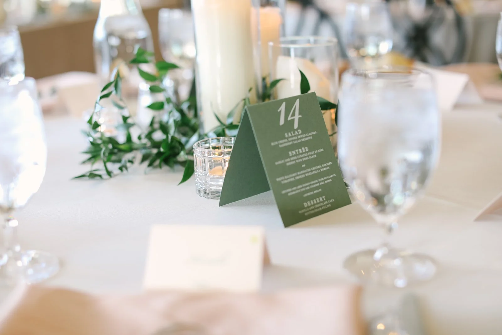 Table setup at an event with a green table card showing the number 14, surrounded by candles, glassware, and greenery in the background.