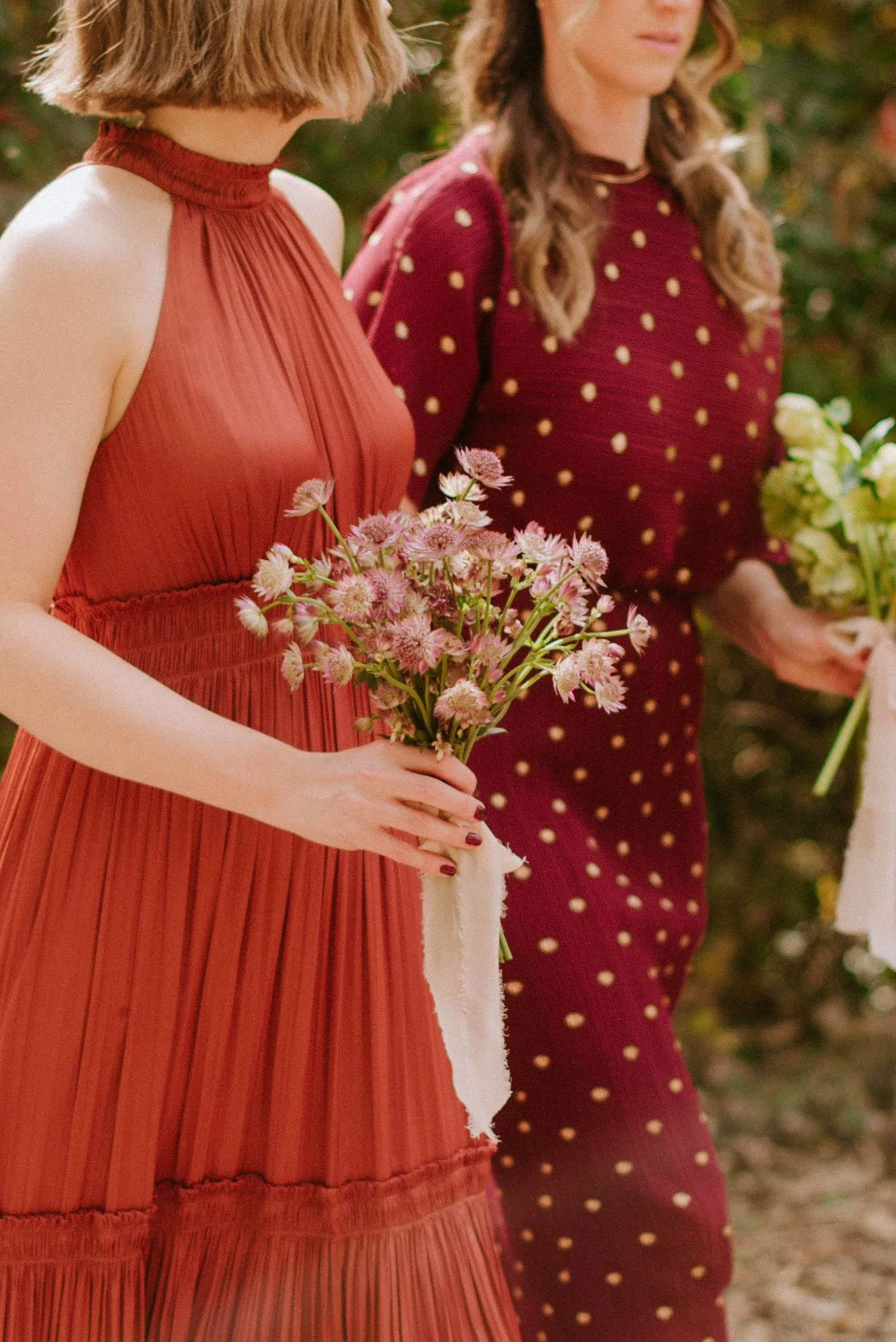 Two women in vintage dresses holding flowers, standing outdoors surrounded by greenery.