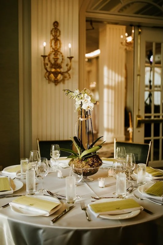 A round dinner table set with glasses, silverware, and yellow menus, featuring a white orchid centerpiece in an elegant room with soft lighting.