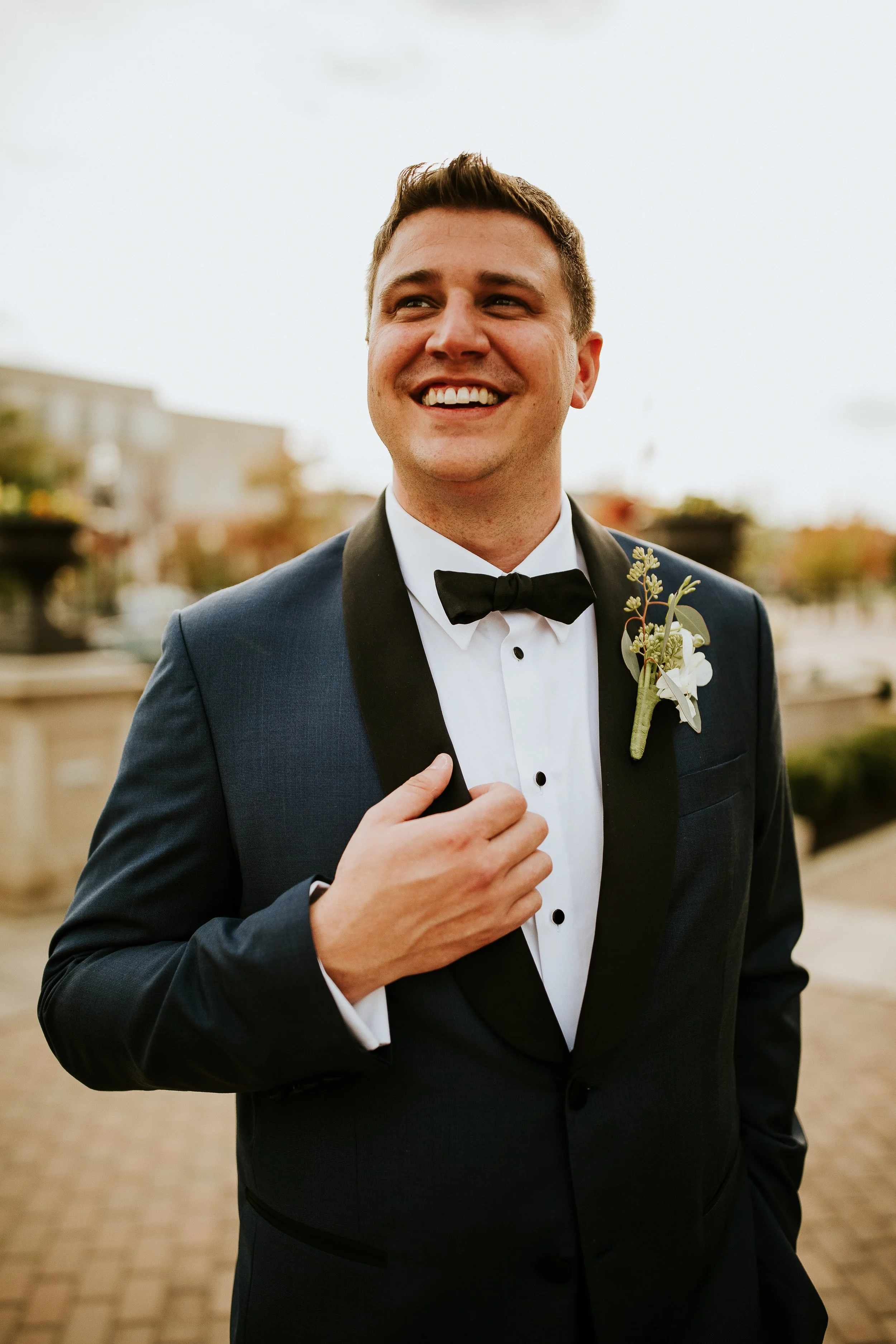 A smiling man in a tuxedo with a bow tie and boutonniere, standing outdoors on a cloudy day.