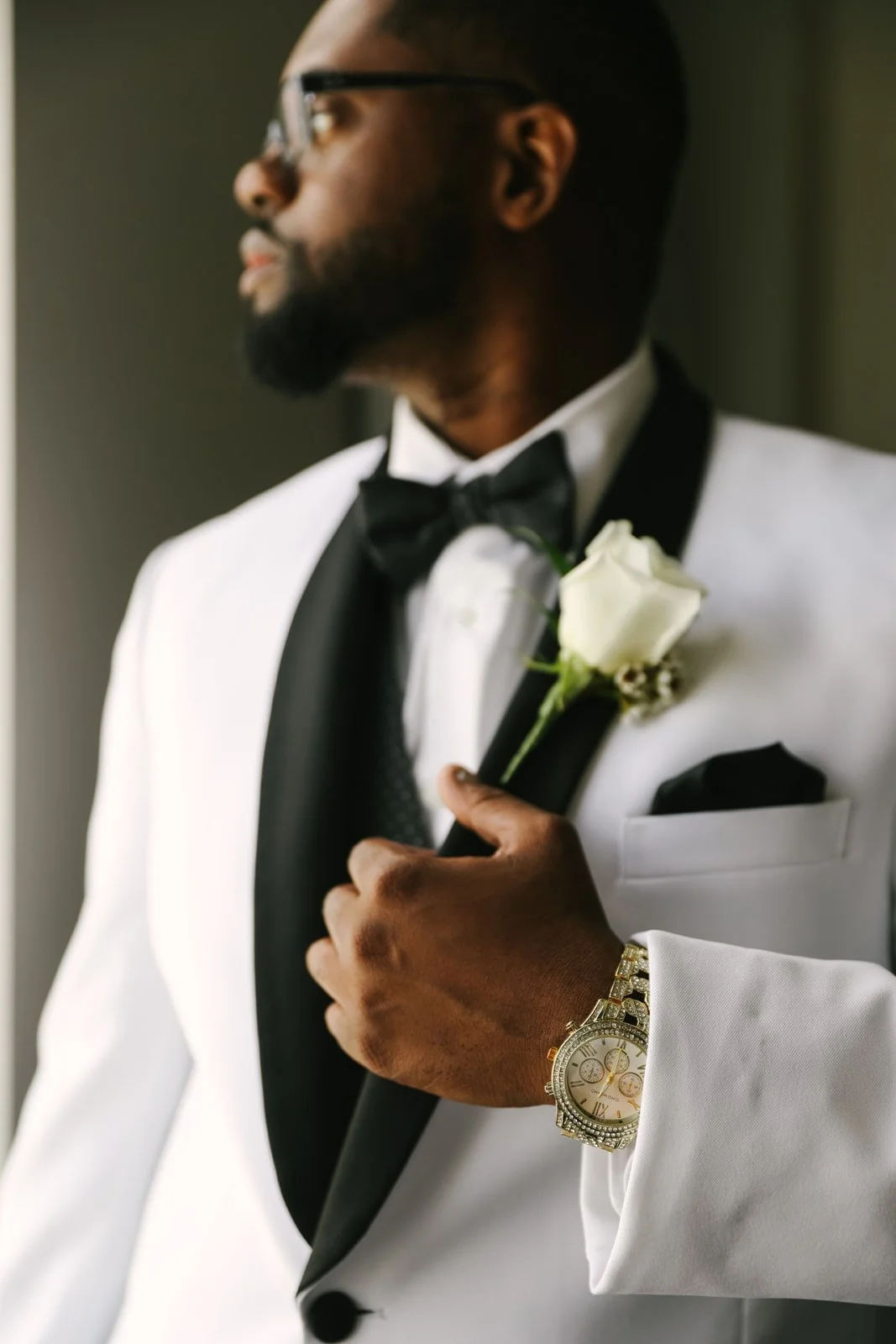 A groom in a white tuxedo with black lapels, wearing a black bow tie, and a gold wristwatch, holding a white rose boutonniere.