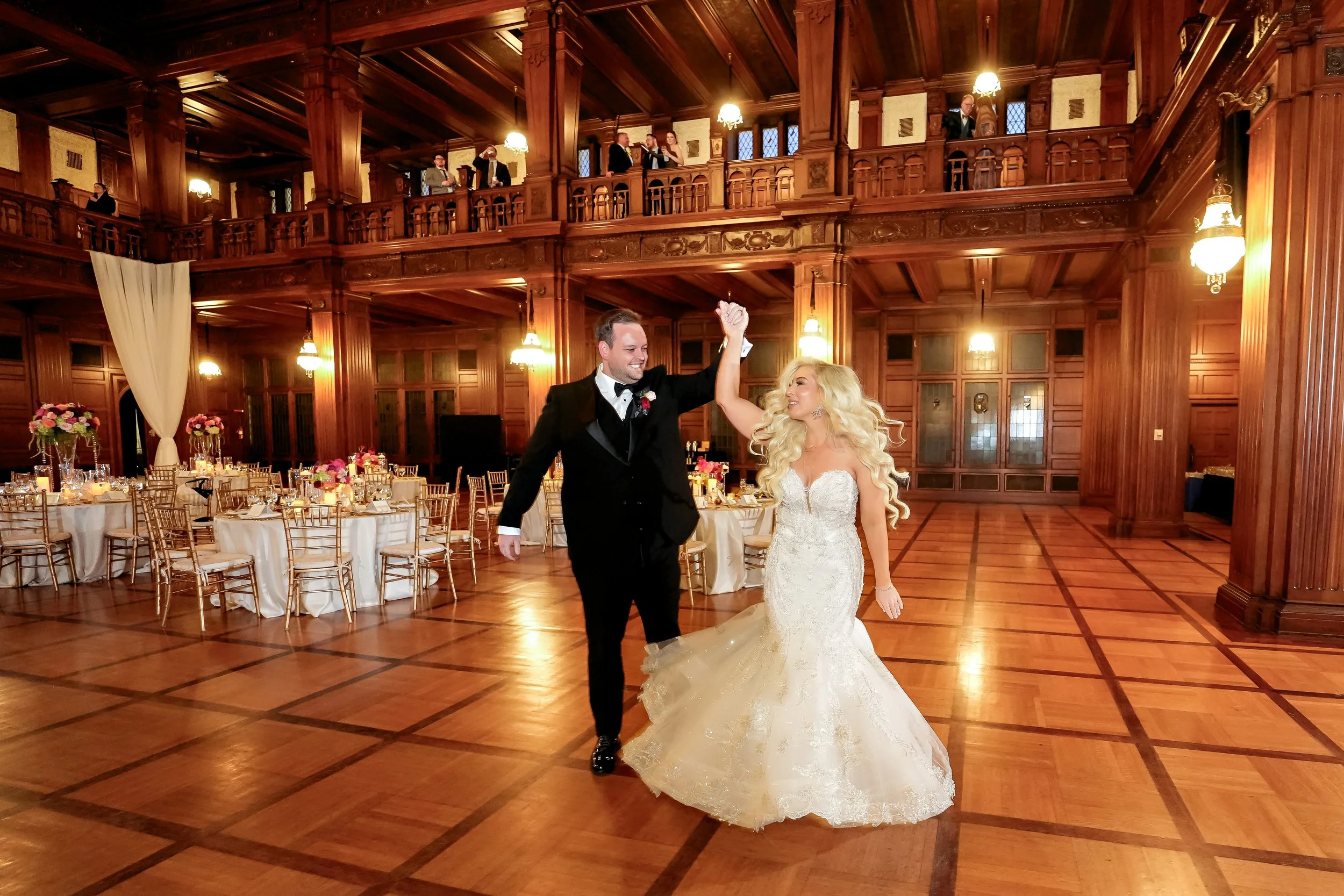A bride and groom dancing at their wedding reception in a grand wooden hall with high ceilings and balconies. The groom is wearing a black tuxedo with a bow tie, and the bride is in a strapless white wedding gown with a full skirt. Guests are seen in
