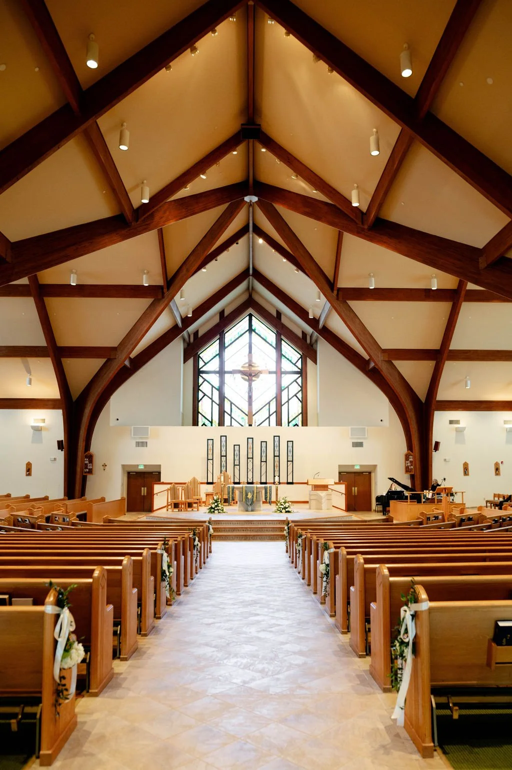 Inside a church with wooden pews decorated with white ribbons and flowers, an altar with flowers, and large stained glass windows behind it.