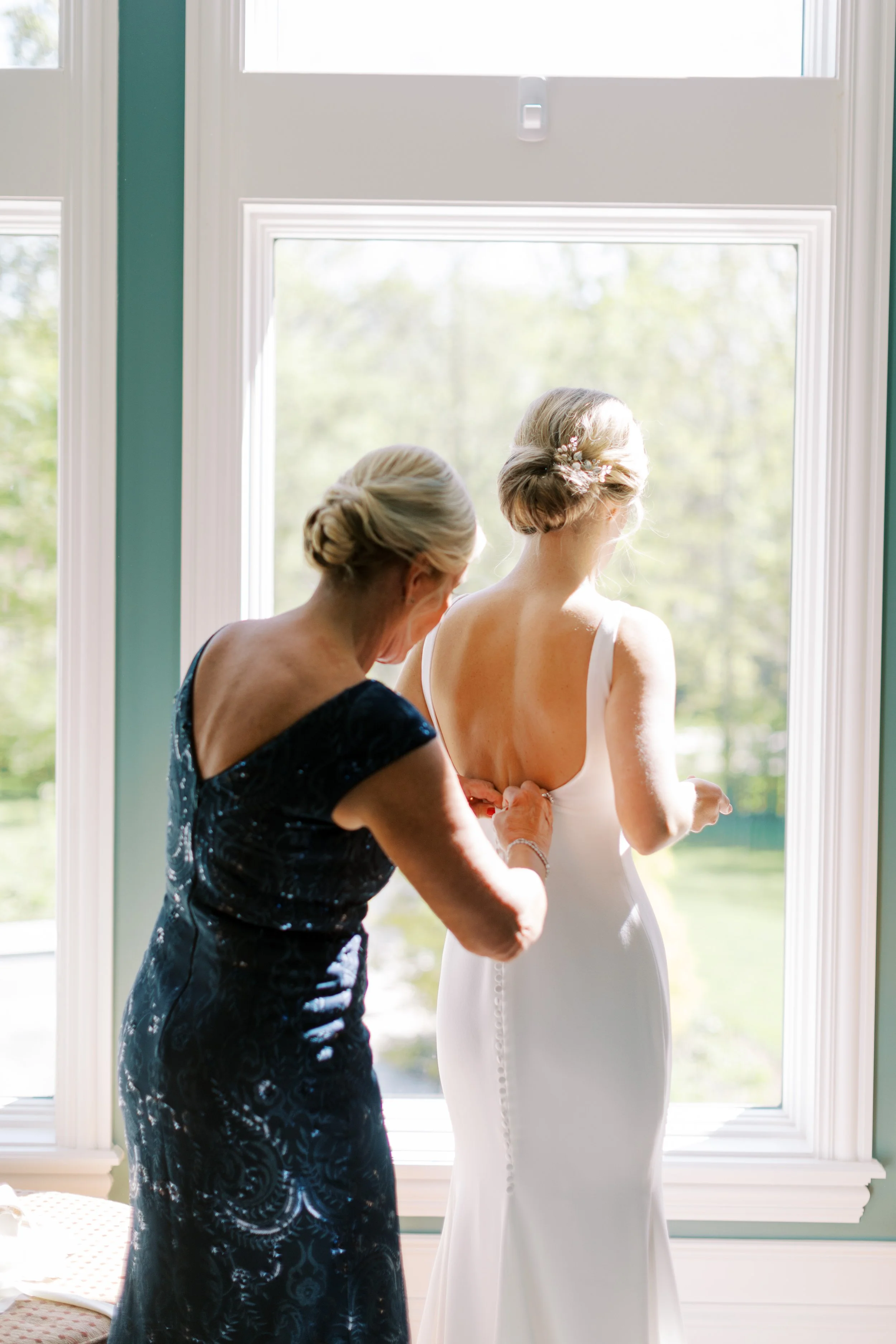 A woman in a black dress helping a bride in a white wedding dress get ready by fastening the back of her dress near a large window.