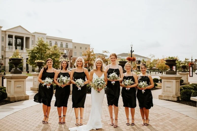 Bridal party of seven women standing outdoors, with the bride in a white dress and six bridesmaids in black dresses, holding white bouquets, in a landscaped area with buildings and trees in the background.