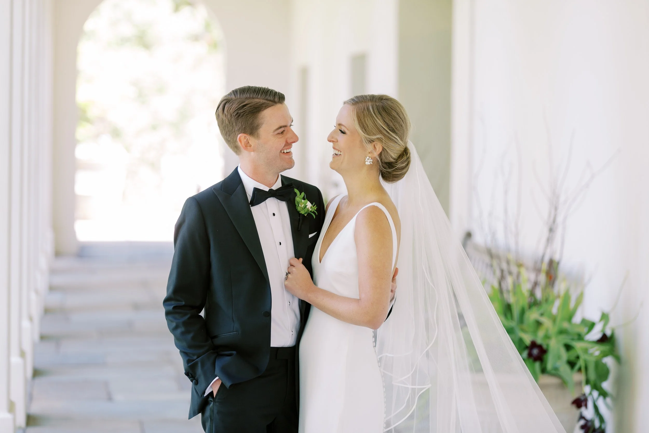 A smiling groom in a tuxedo and a bride in a white wedding dress and veil facing each other and smiling inside a bright, airy space with white walls and stairs in the background.