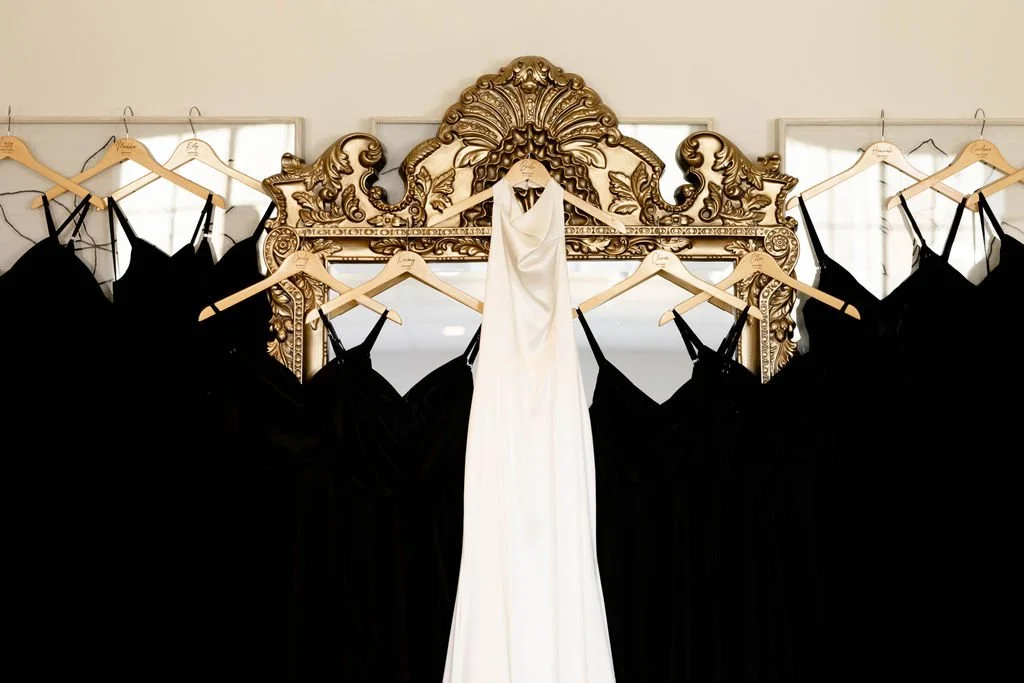 A white wedding dress hanging on a hanger in the center with black dresses on either side, all on wooden hangers, in front of an ornate gold mirror.