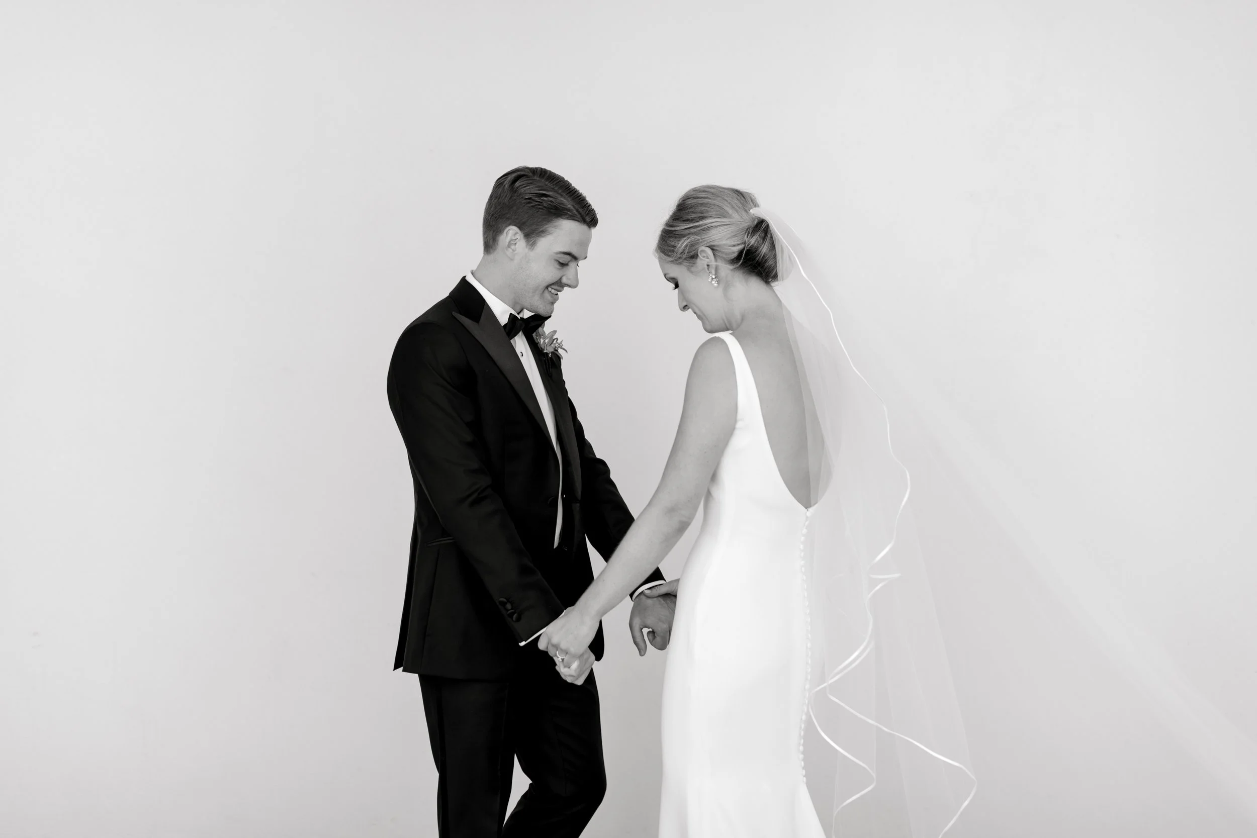 A black and white photo of a bride and groom holding hands, smiling and looking at each other, standing against a plain background.
