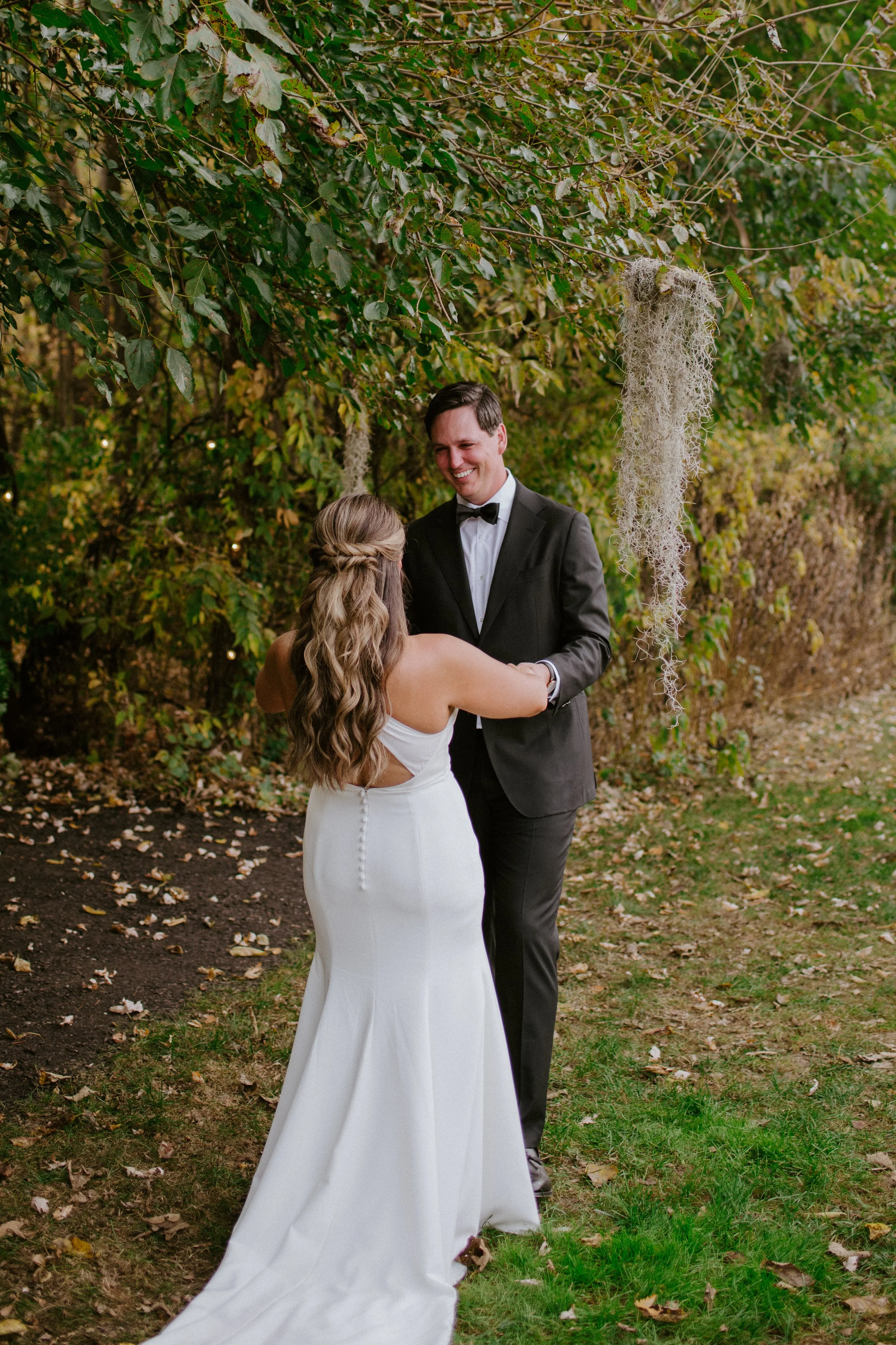 A bride and groom are dancing outdoors under a tree decorated with Spanish moss, with the bride wearing a white gown and the groom in a black tuxedo, surrounded by fallen leaves.