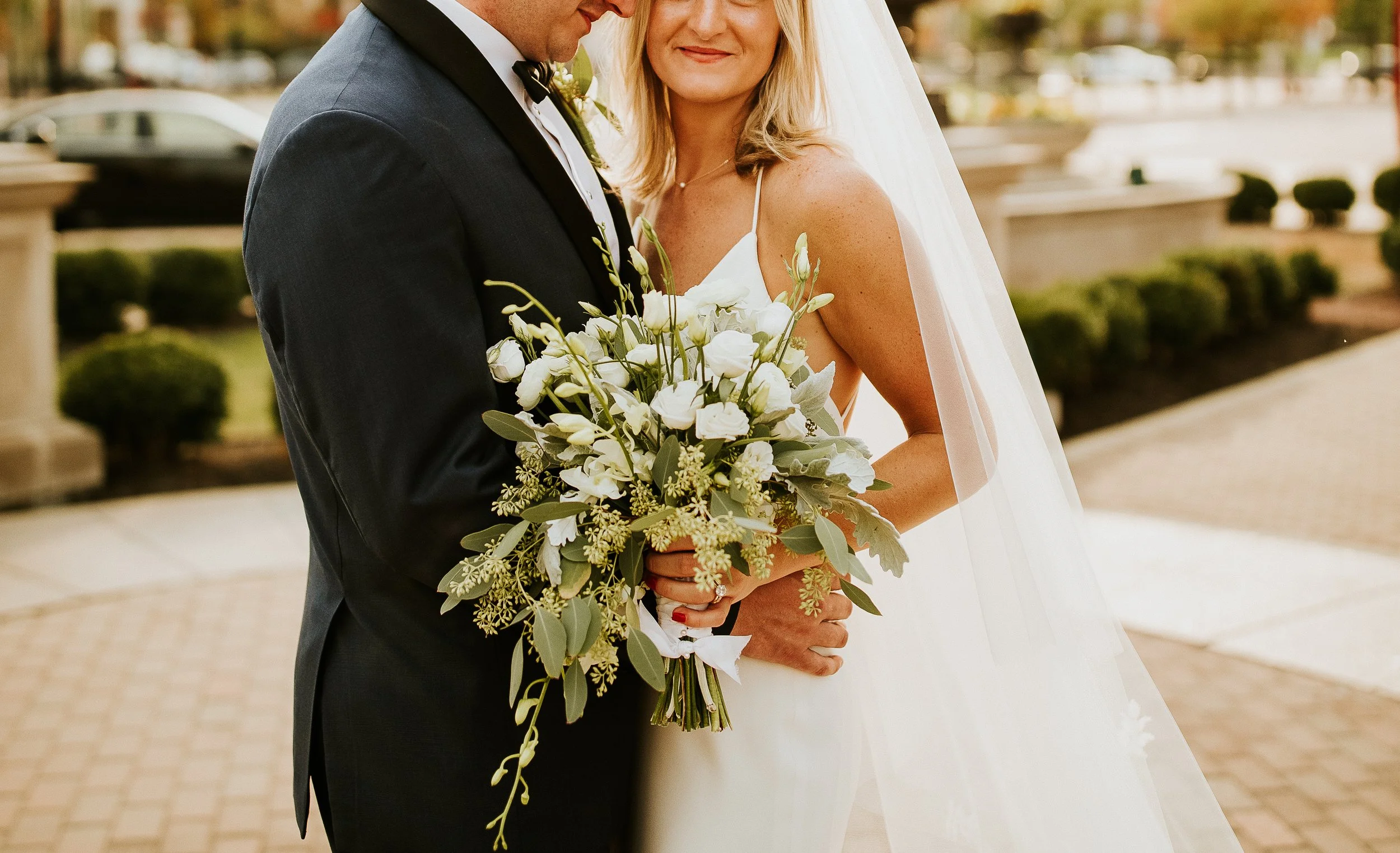 Bride and groom standing close together, with the bride holding a bouquet of white flowers and greenery. The groom is wearing a black tuxedo with a bow tie, and the bride is wearing a white wedding dress with thin straps and a veil. They are outdoors