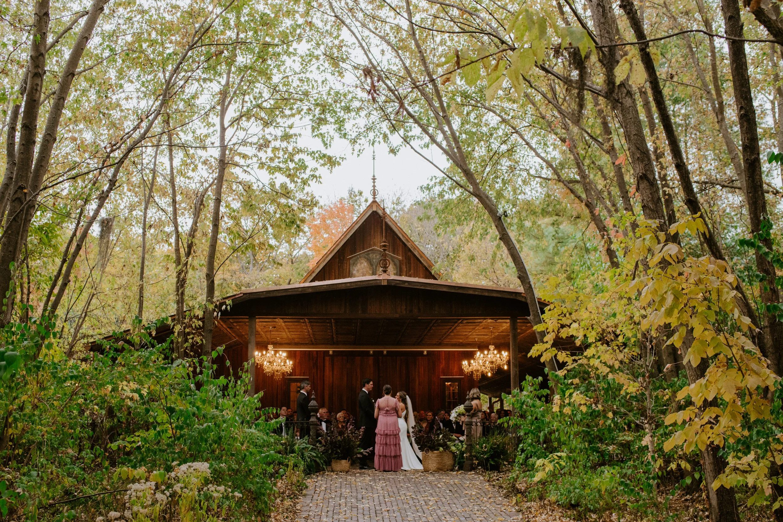 Wedding ceremony taking place inside a rustic barn surrounded by trees with autumn leaves, chandeliers hanging from the ceiling, and guests seated. The bride and groom are standing at the altar, facing each other.