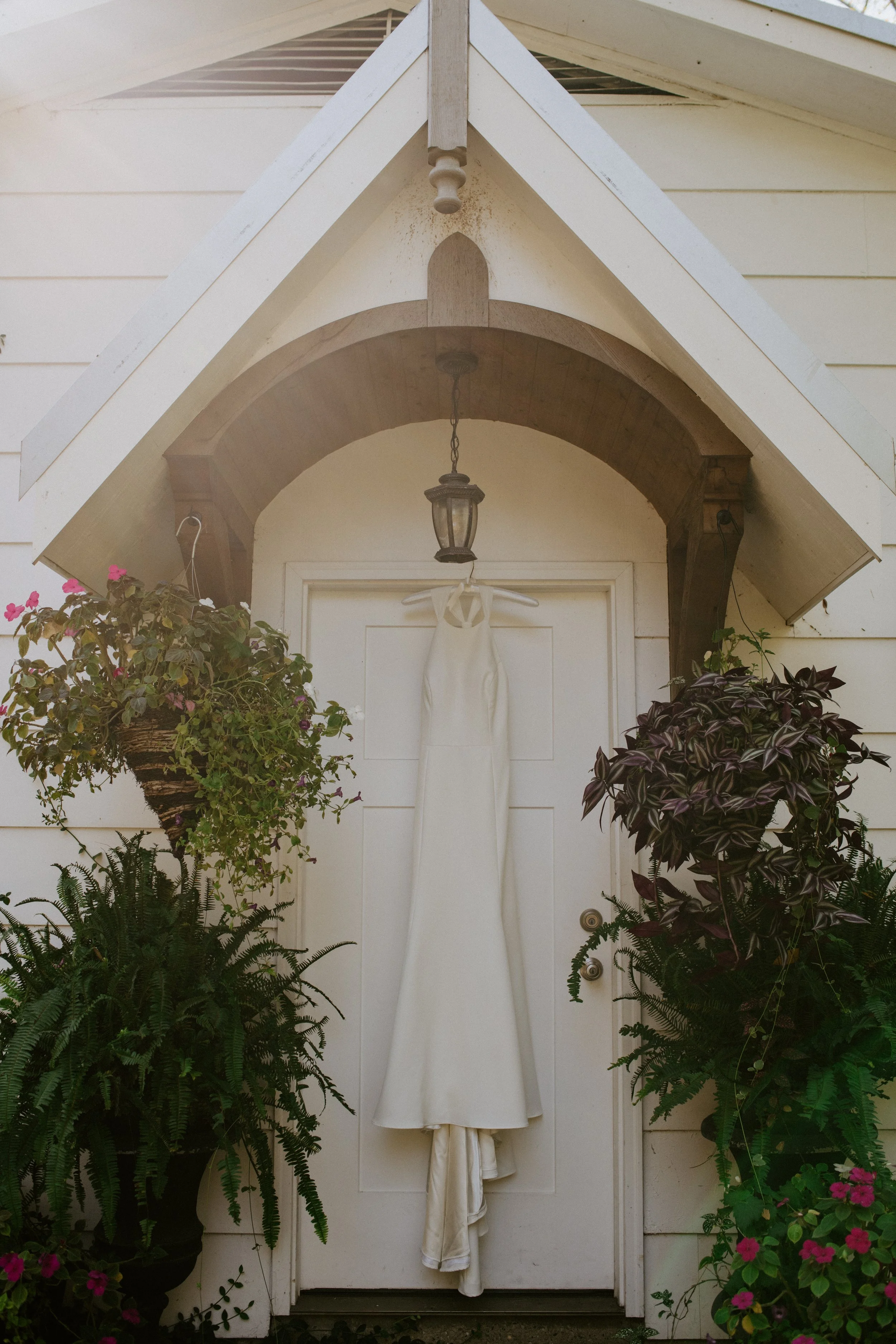 A white door with a wedding dress hanging on a hanger attached to a hook on the door. The door is framed by an archway with plants on either side, including ferns and flowering plants. There is a light fixture hanging above the door under a small woo
