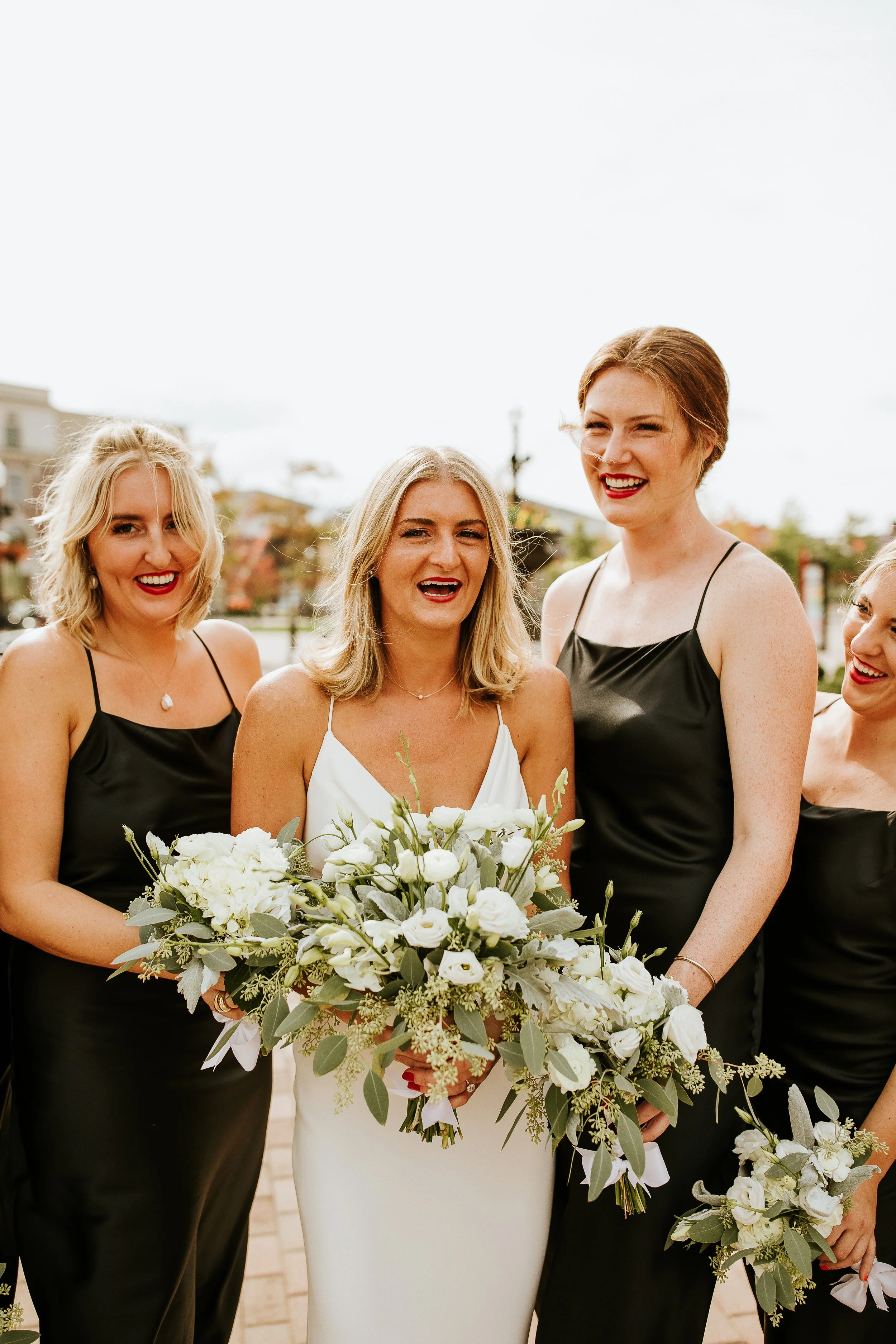 Group of women in formal attire at a wedding, holding bouquets of white flowers, smiling and standing outdoors.