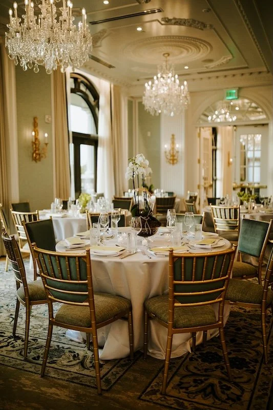 Elegant banquet hall with chandeliers, round tables set with white tablecloths, glassware, and flowers, in a finely decorated room with large windows and ornate detailing.