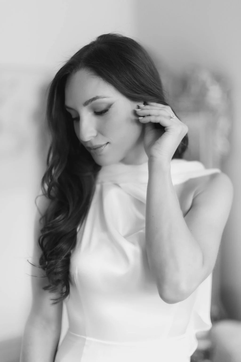 Black and white photo of a woman with long wavy hair, dressed in a sleeveless satin dress, touching her earring with her eyes closed, standing against a plain background.