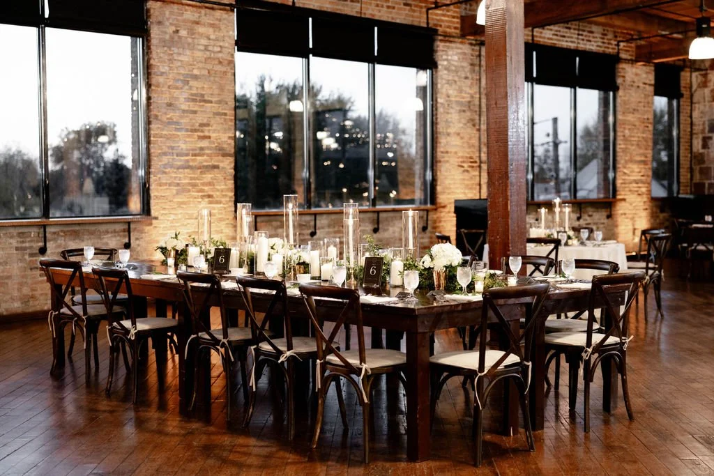 A decorated banquet table set with white flowers, candles, table numbers, and glassware indoors at an event space with large windows and brick walls.