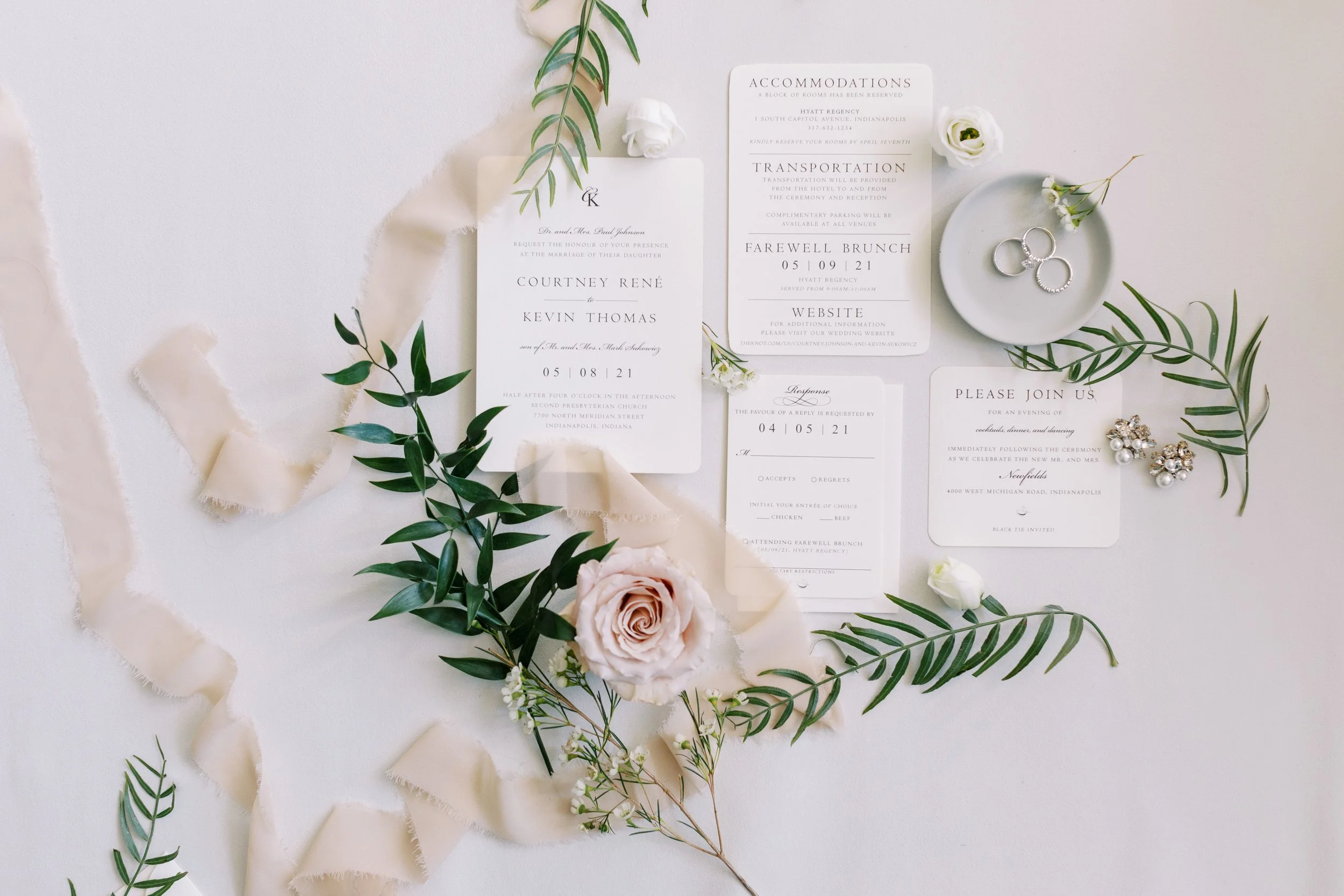 Wedding invitations, floral decorations, rings on a small dish, and loose greenery and flowers on a white surface.