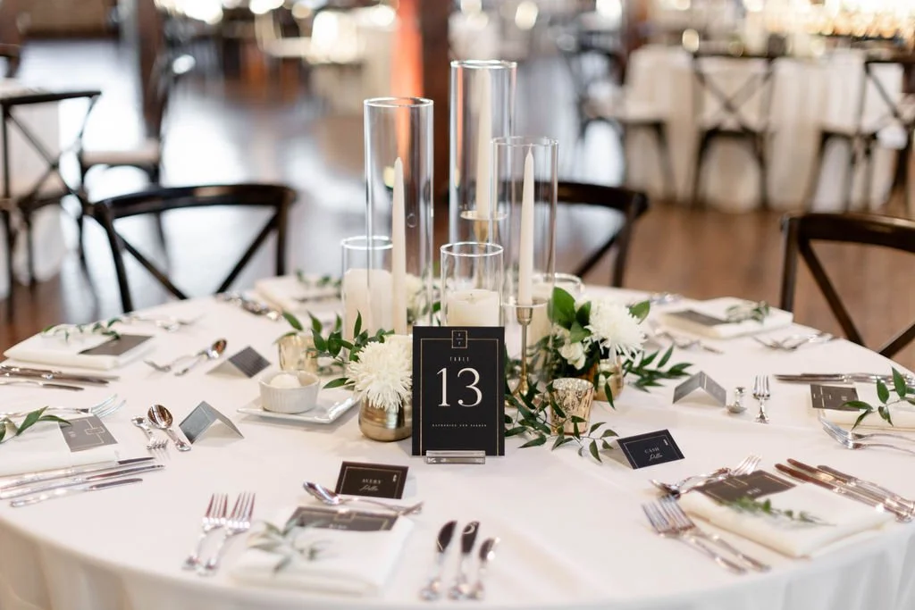 A round wedding reception table decorated with white flowers, greenery, and tall glass candle holders with white candles. The table is set with silverware, plates, and folded napkins with sprigs of greenery. A black table number sign displays the num