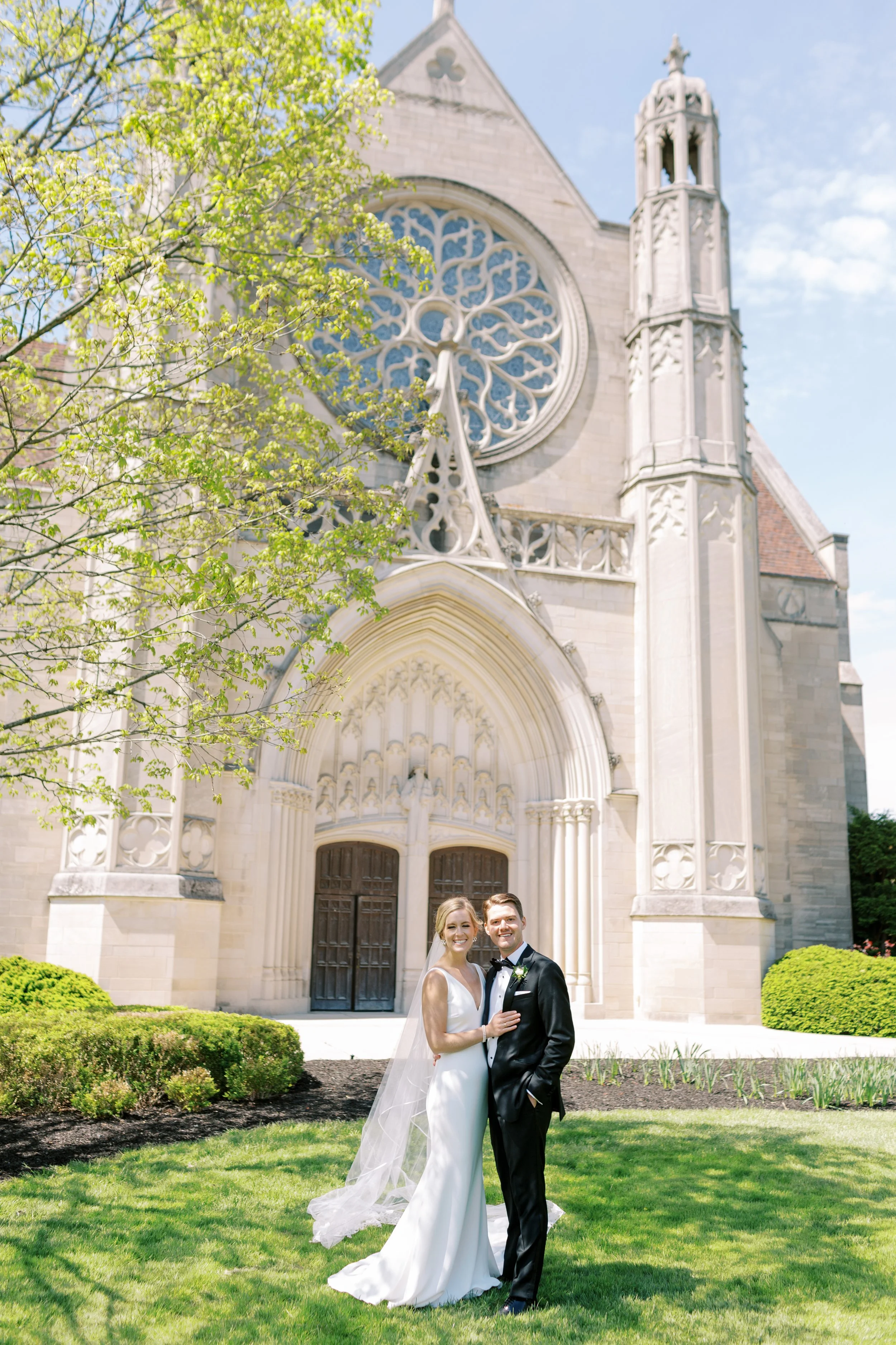 A bride and groom in wedding attire standing outdoors on a lawn in front of a large church with stained glass windows and ornate architecture. The bride is wearing a white dress and veil, and the groom is in a black tuxedo. There are lush bushes and 