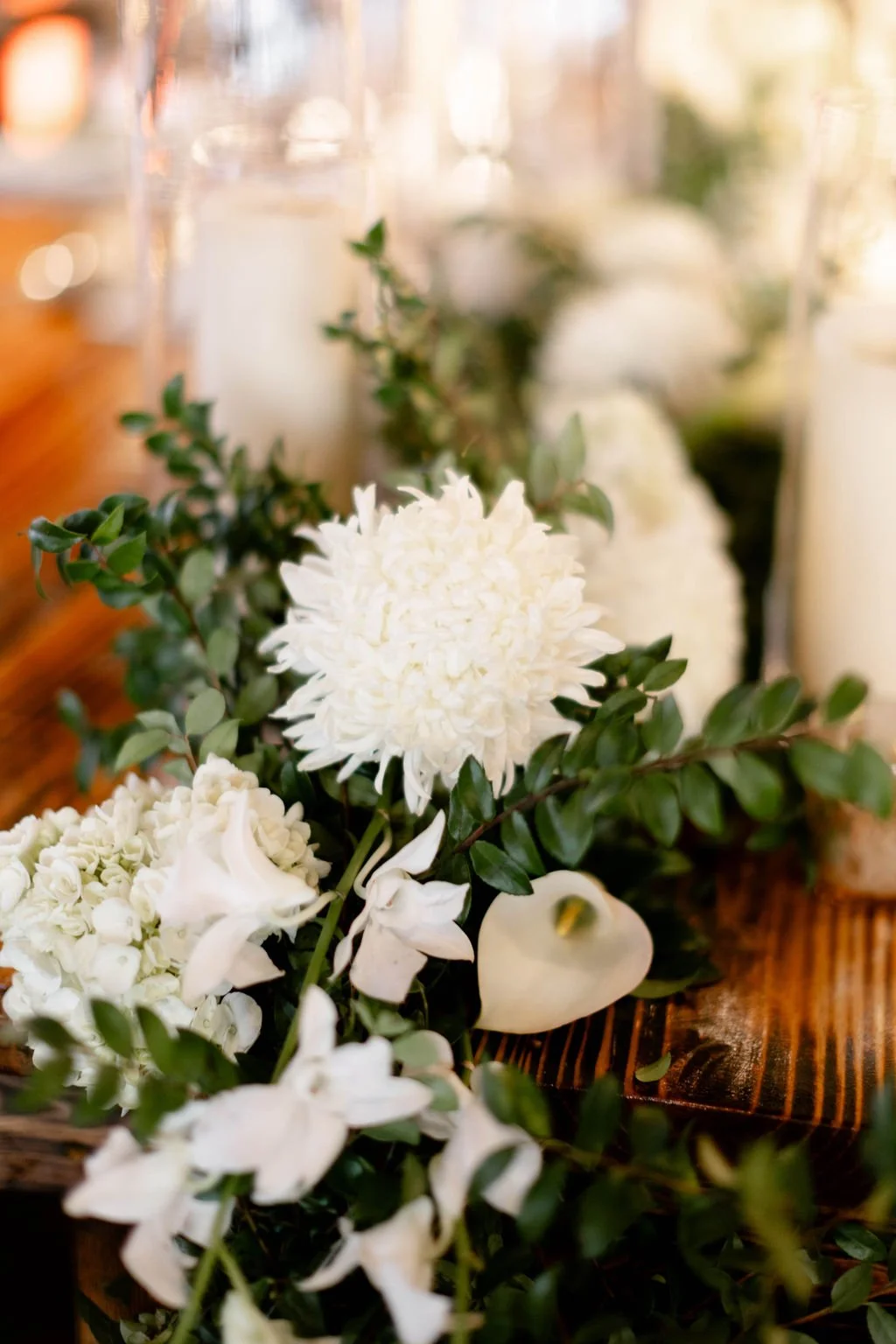 Close-up of a white floral arrangement with green leaves on a wooden table, blurred candles in the background.
