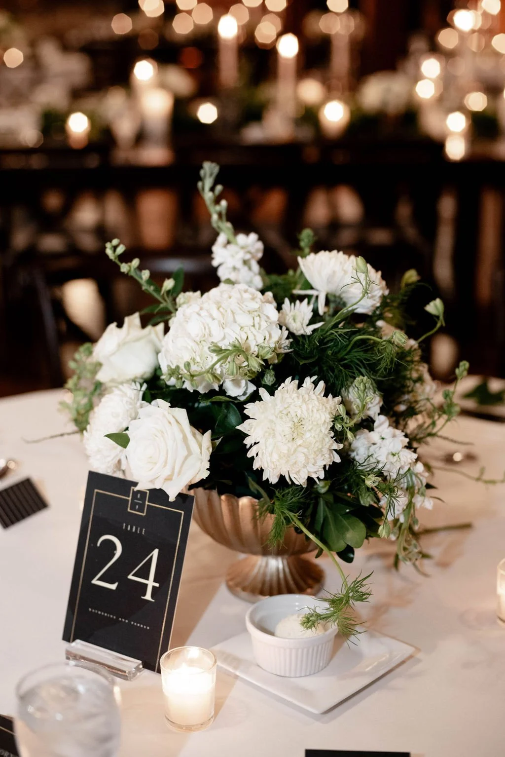A floral centerpiece with white flowers in a copper bowl on a table at a formal event, with table number 24 sign, candles, and a white tablecloth in the dimly lit background.