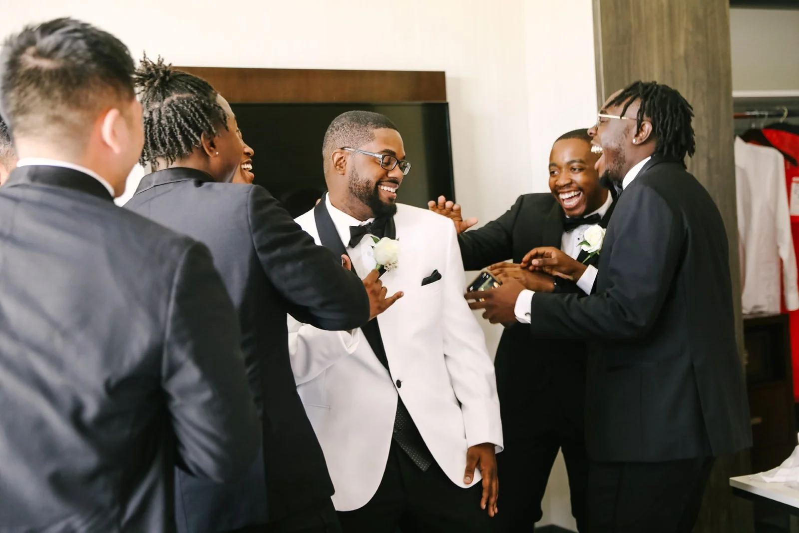 A group of men in tuxedos, laughing and celebrating in a room, with one man wearing a white tuxedo jacket.