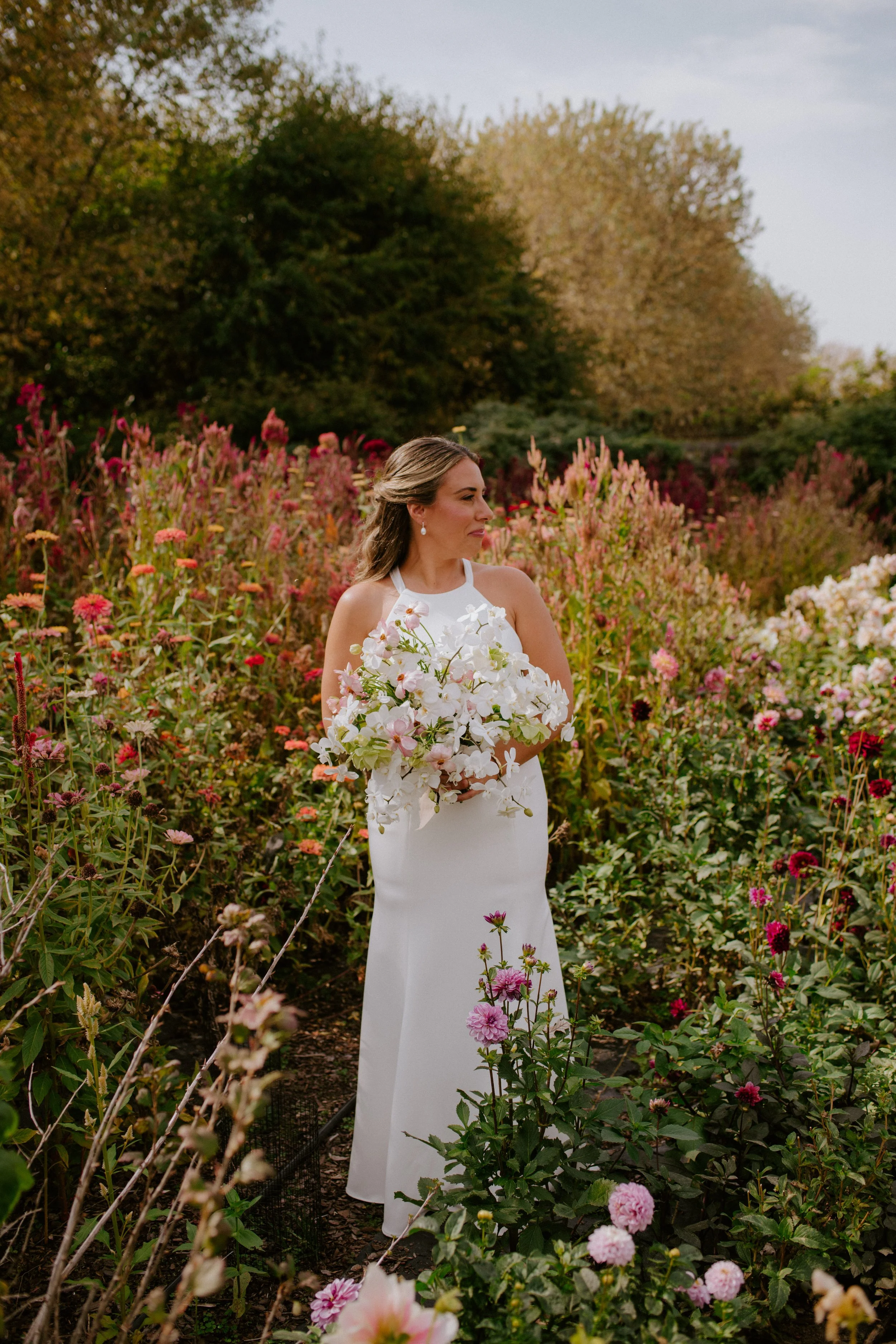 A woman in a white dress holding a bouquet of white flowers standing in a garden with pink, white, and purple flowers and trees in the background.