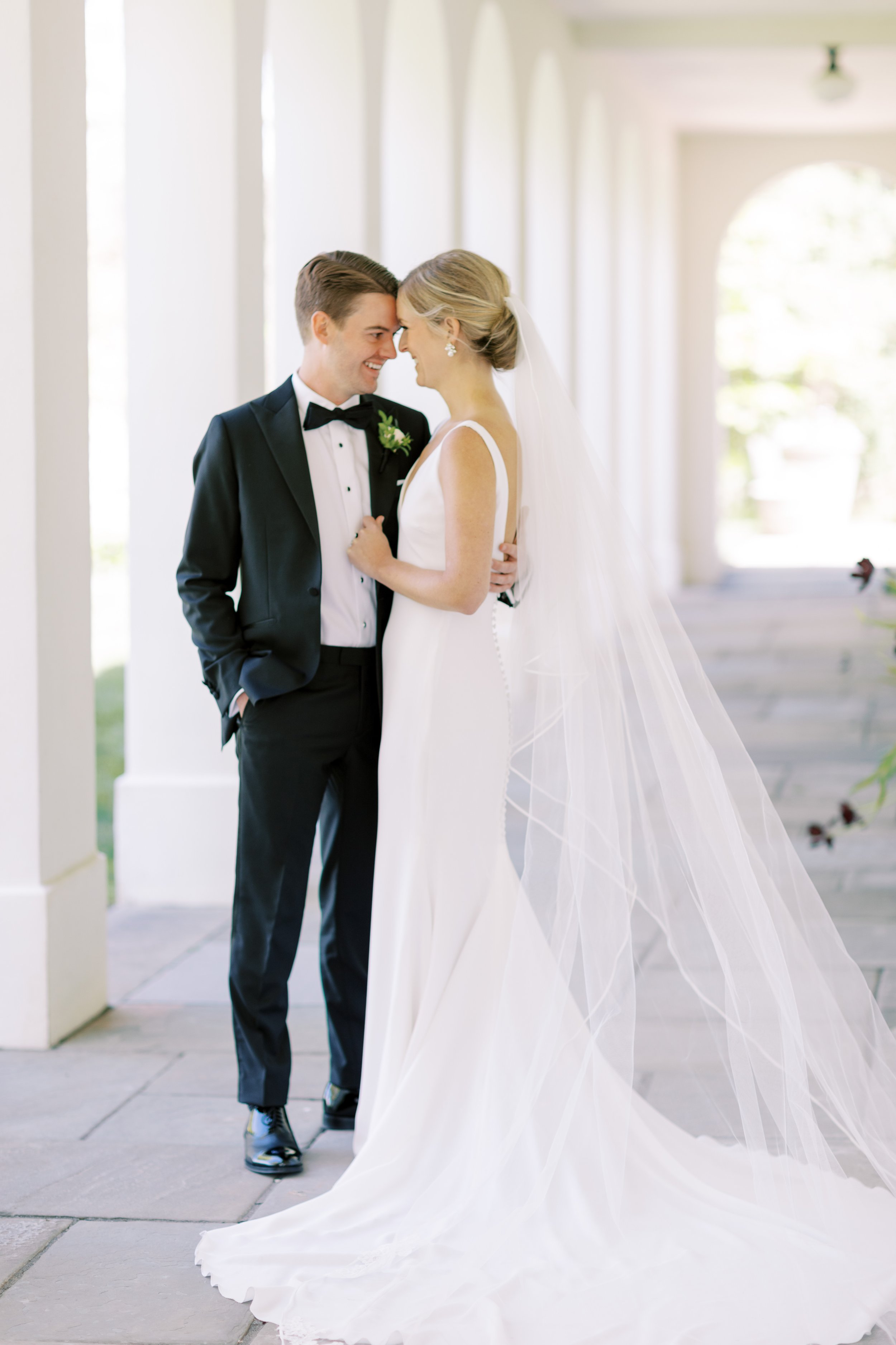 A bride and groom stand closely facing each other, smiling, under a series of white arches with natural light streaming through. The bride wears a white wedding gown with a long veil, and the groom wears a black tuxedo with a bow tie.
