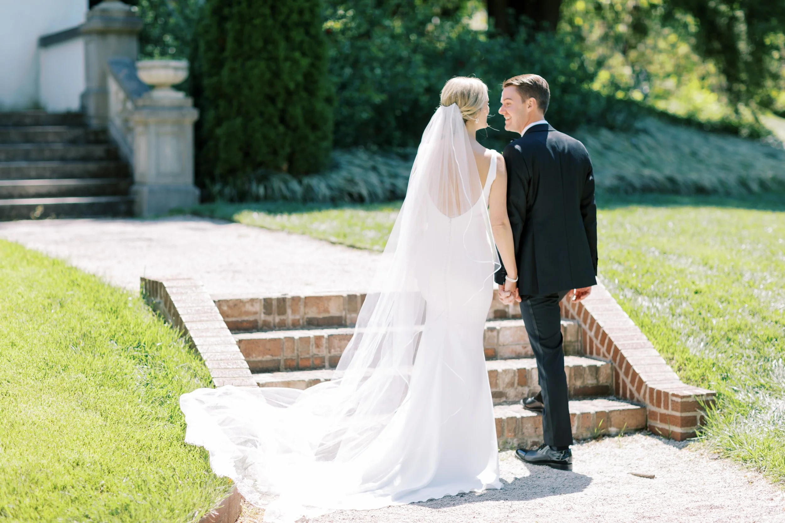 A bride in a white wedding dress with a train and veil holding hands with a groom in a black suit outside near brick steps and greenery, smiling at each other.