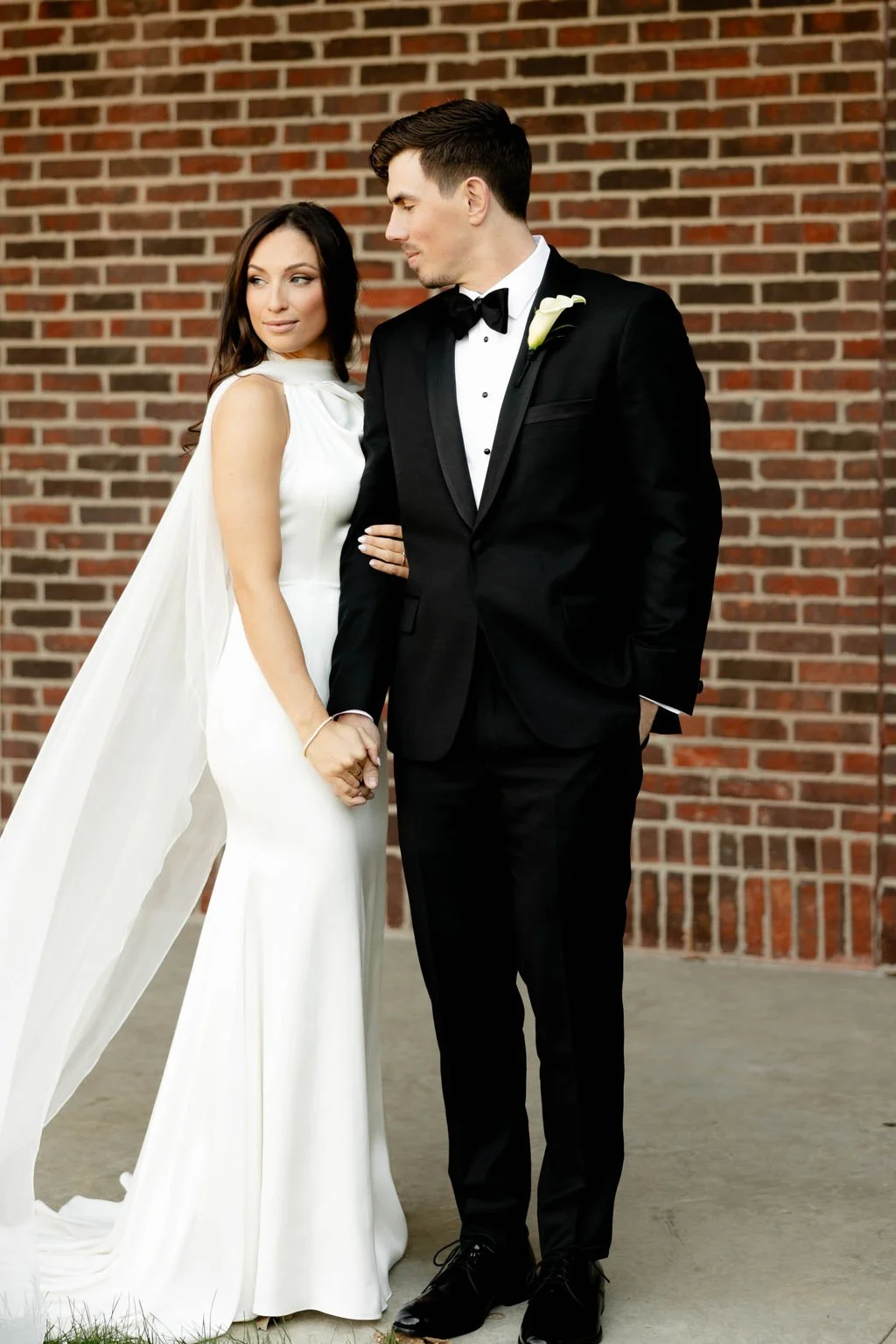 A bride and groom holding hands, standing in front of a brick wall. The bride is wearing a white wedding dress with a cape, and the groom is dressed in a black tuxedo with a bow tie and a white flower boutonniere.