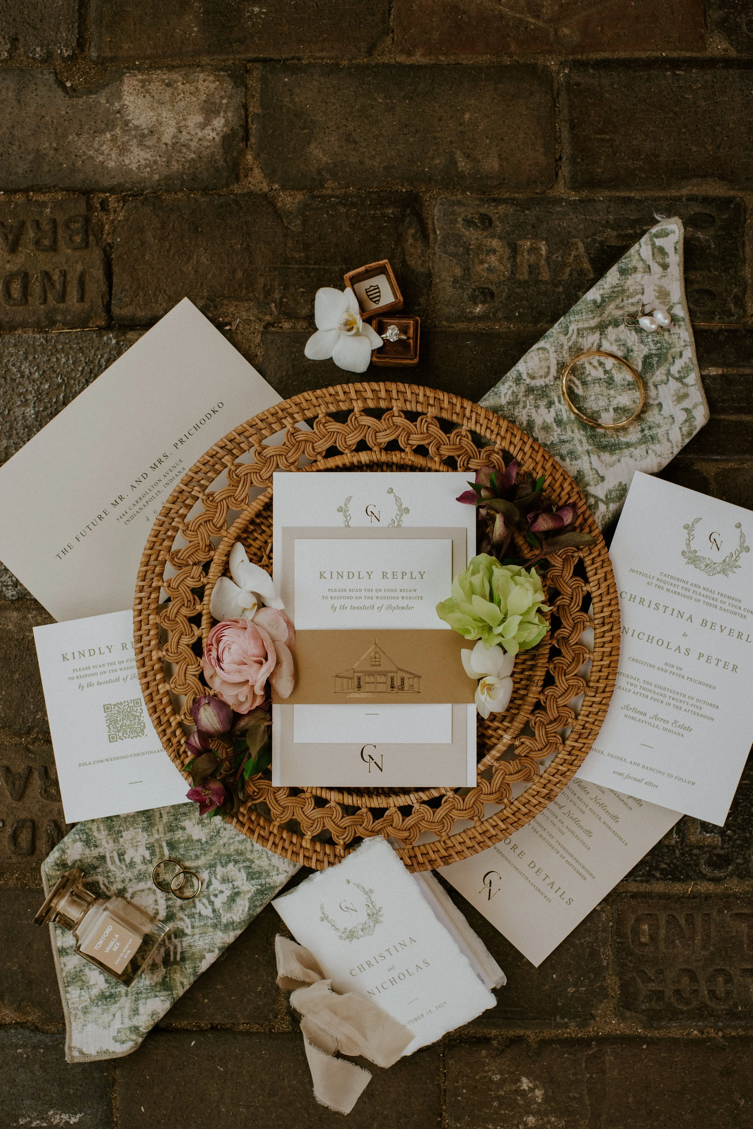 Wedding stationery and accessories arranged on a rustic brick surface. Items include invitations with elegant fonts, a woven basket with flowers, an open jewelry box with a ring, a gold bracelet, and pearl earrings on a fabric pouch.