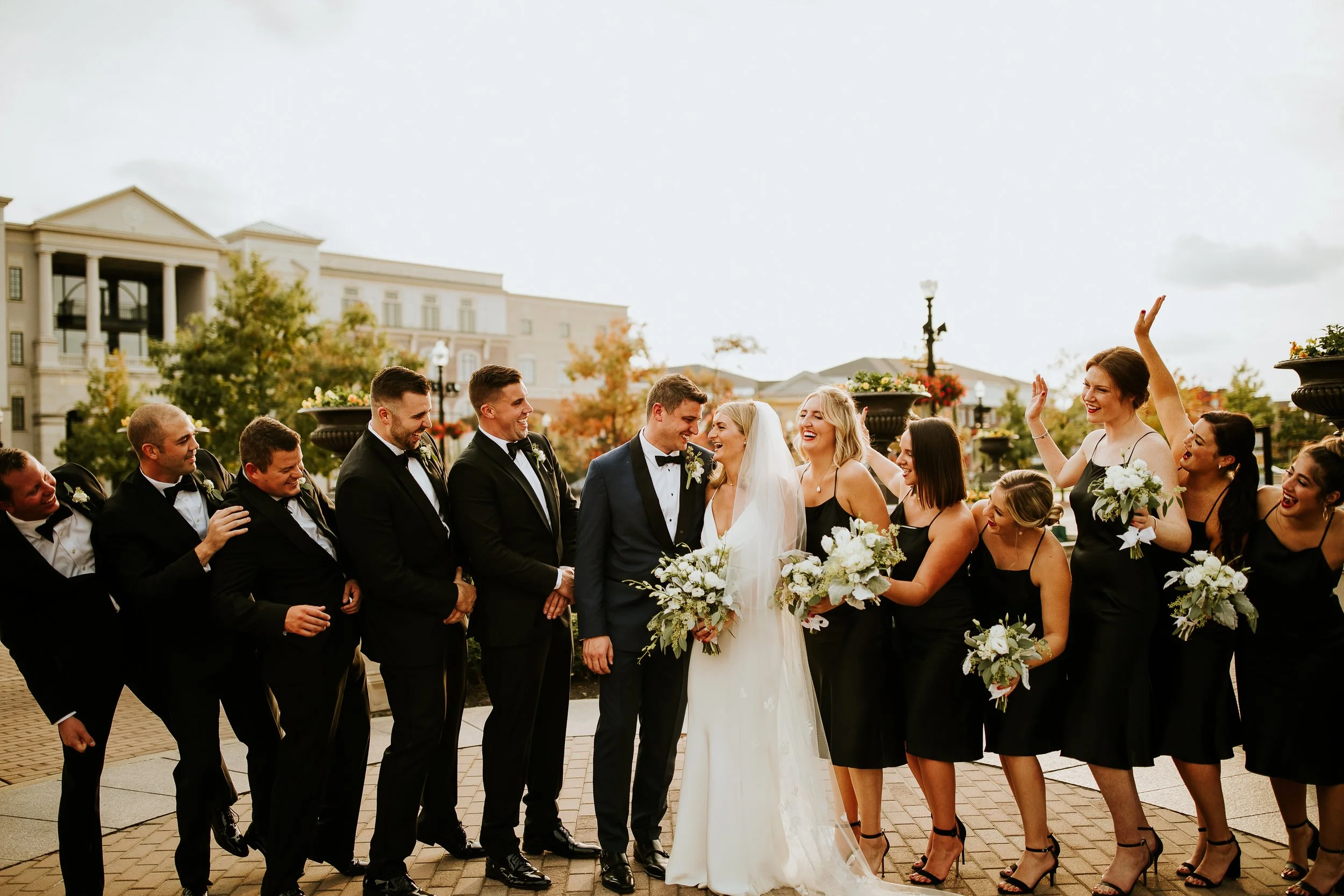 A group of wedding guests, including the bride and groom, standing outdoors in front of a building during a wedding celebration. The bride in a white dress and veil is holding a bouquet of flowers, and the groom is dressed in a tuxedo. The guests are