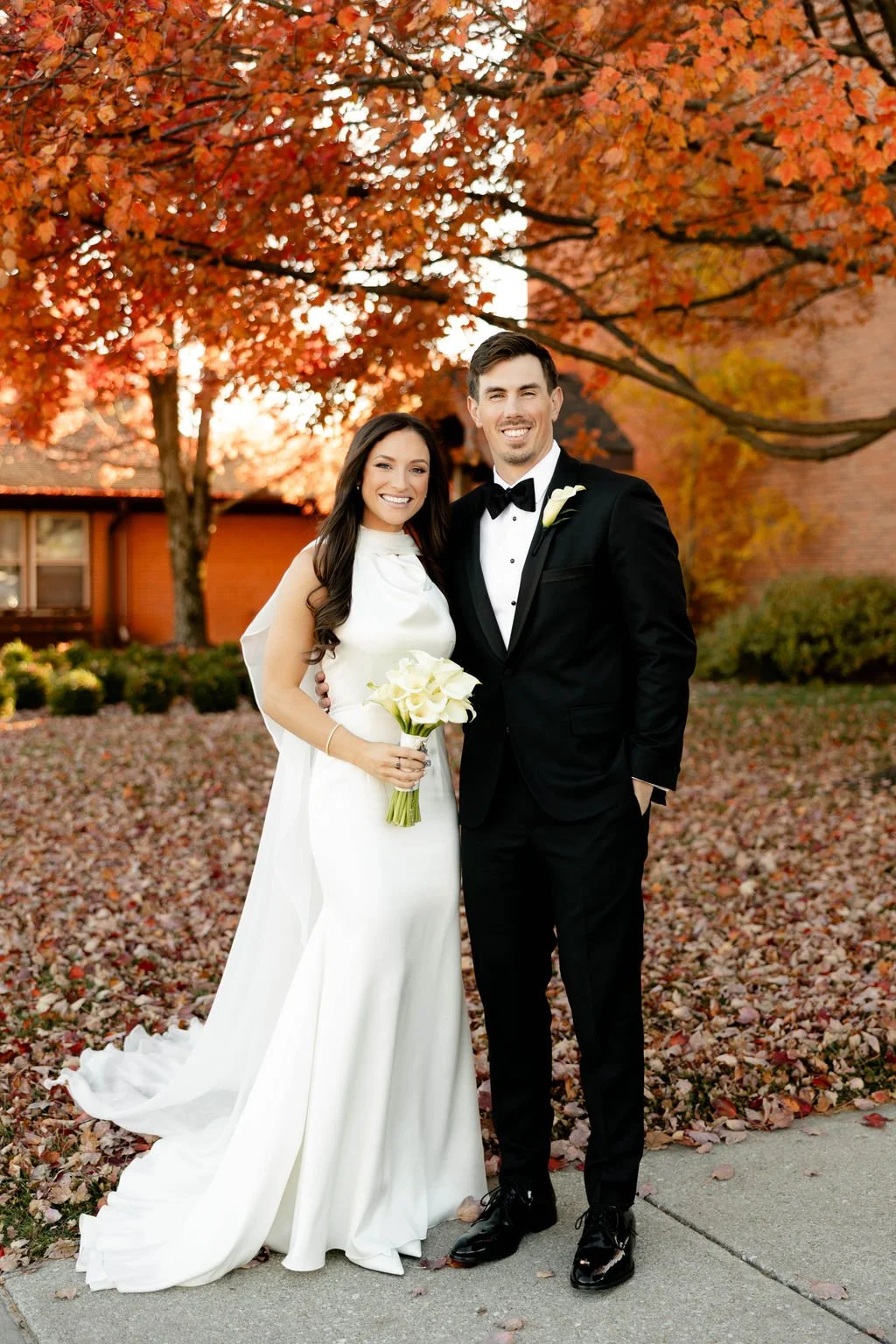 A bride and groom smiling and standing close together outdoors during autumn, with colorful fall foliage and a house in the background. The bride wears a white gown and holds a bouquet of white flowers, while the groom is in a black tuxedo with a bow