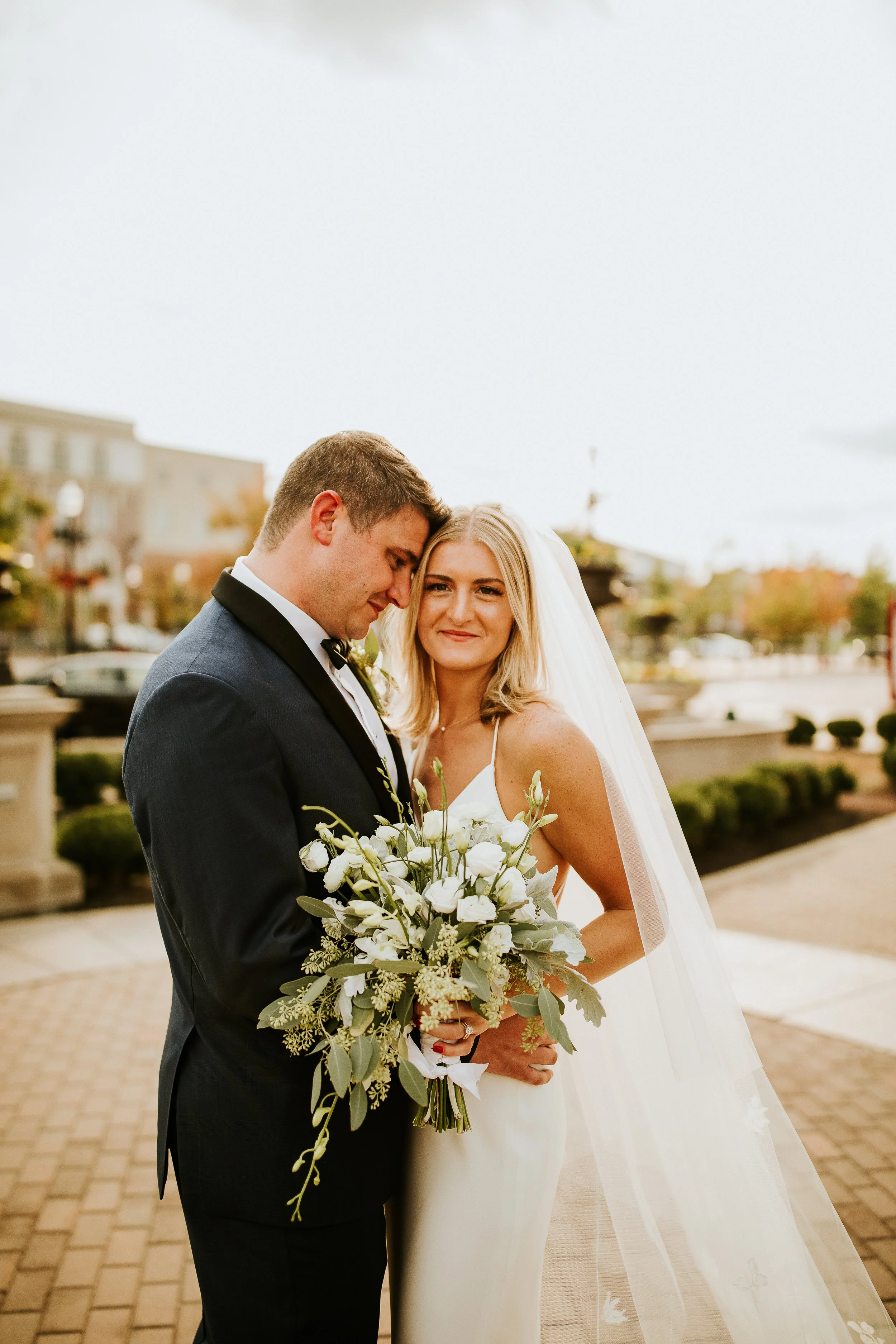 A bride and groom stand close together outdoors, with the groom's head resting against the bride's, she smiling gently and holding a bouquet of white flowers and greenery, in front of a background of buildings, trees, and a cloudy sky.