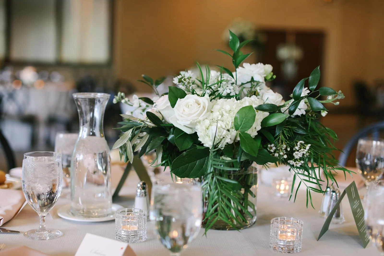 A floral centerpiece with white roses, lilies, and greenery on a table set for a formal event, with glasses, candles, and table cards in the background.