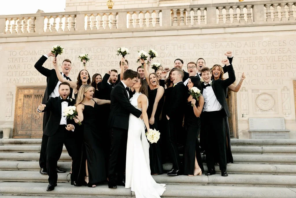 A wedding party on steps, with the bride and groom kissing, others celebrating around them, some holding bouquets and raising their fists in celebration.
