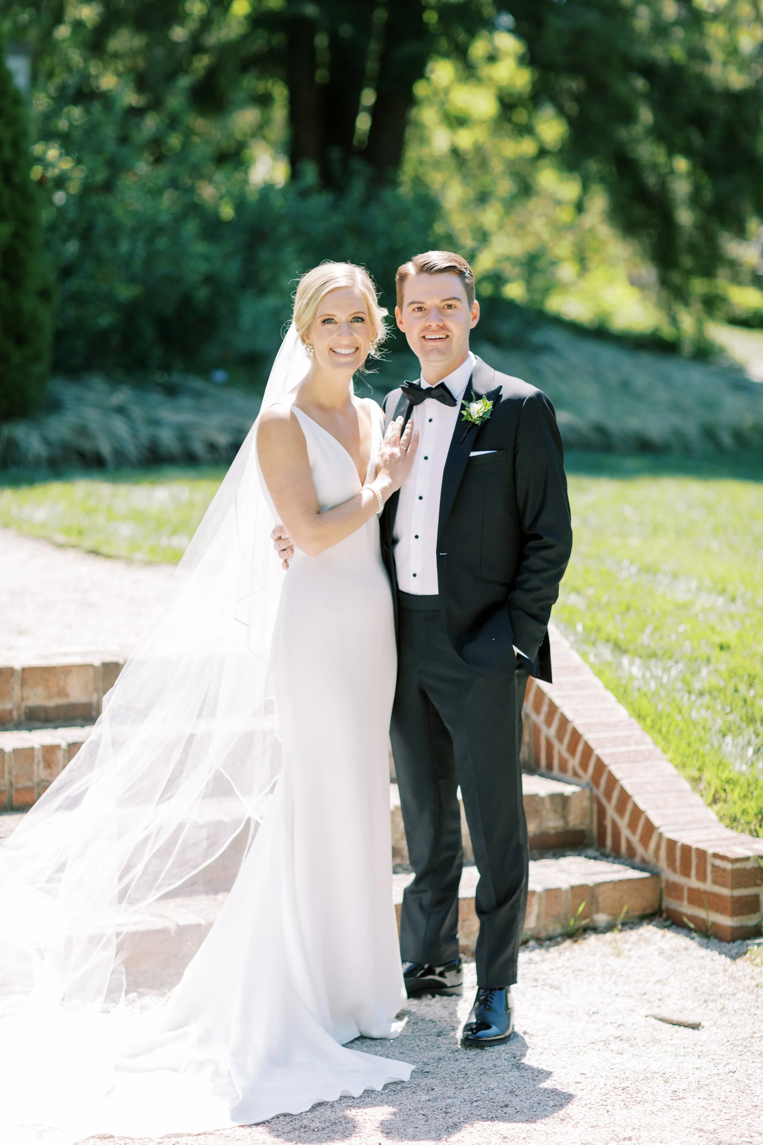 A newlywed couple standing outdoors on a sunny day, with greenery in the background, smiling at the camera. The bride is wearing a white wedding dress with a veil, and the groom is in a black tuxedo with a bow tie and boutonnière.