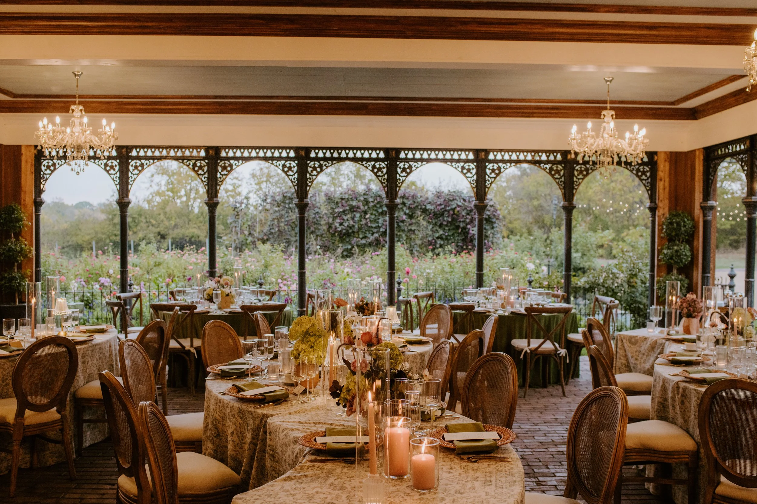 Elegant indoor wedding reception setup with round tables covered with lace tablecloths, floral centerpieces, candles, and chairs, with large arched windows, greenery outside, and chandeliers hanging from the ceiling.