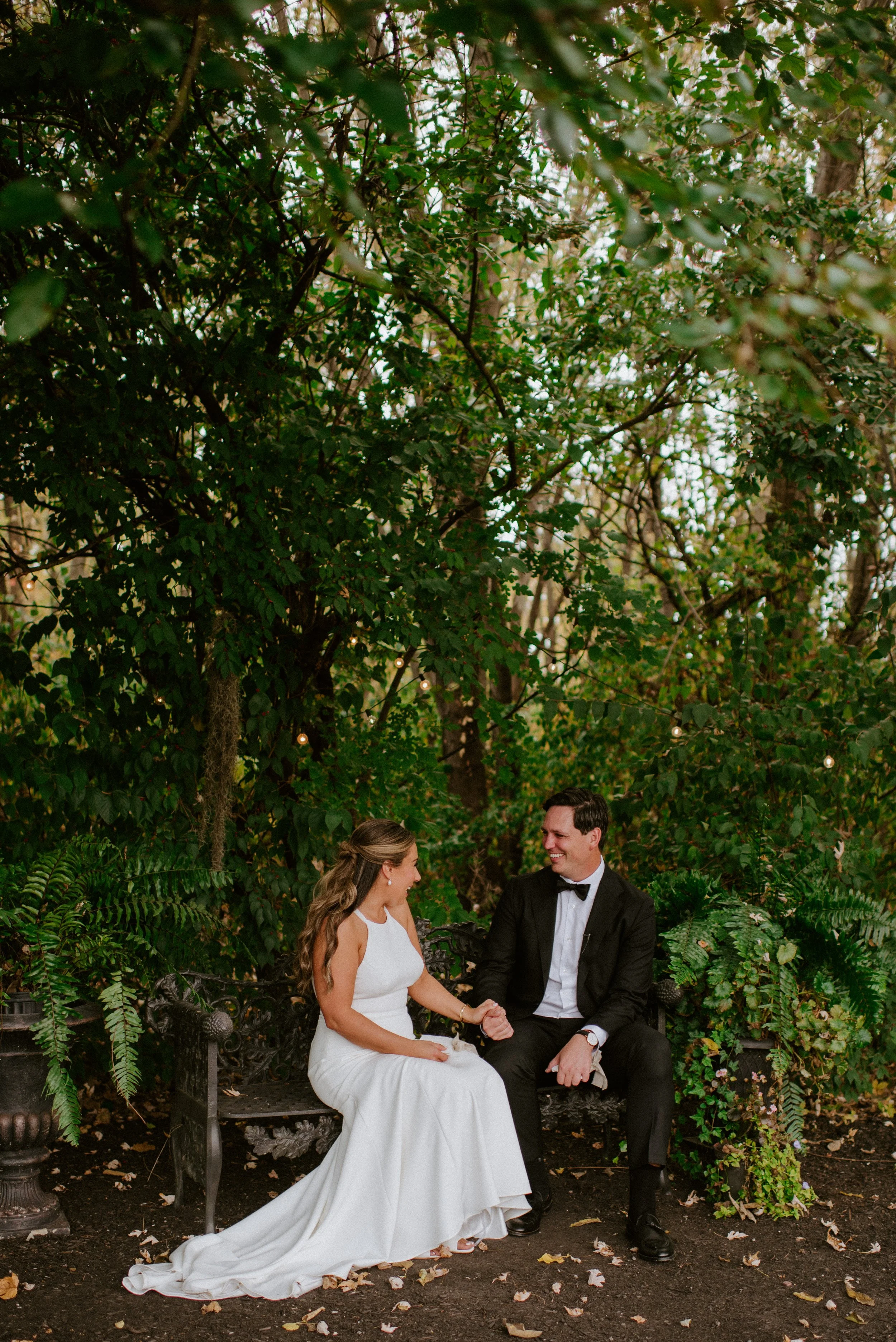 A bride and groom sitting on a bench in a park, holding hands and smiling at each other, surrounded by lush greenery and fallen leaves.