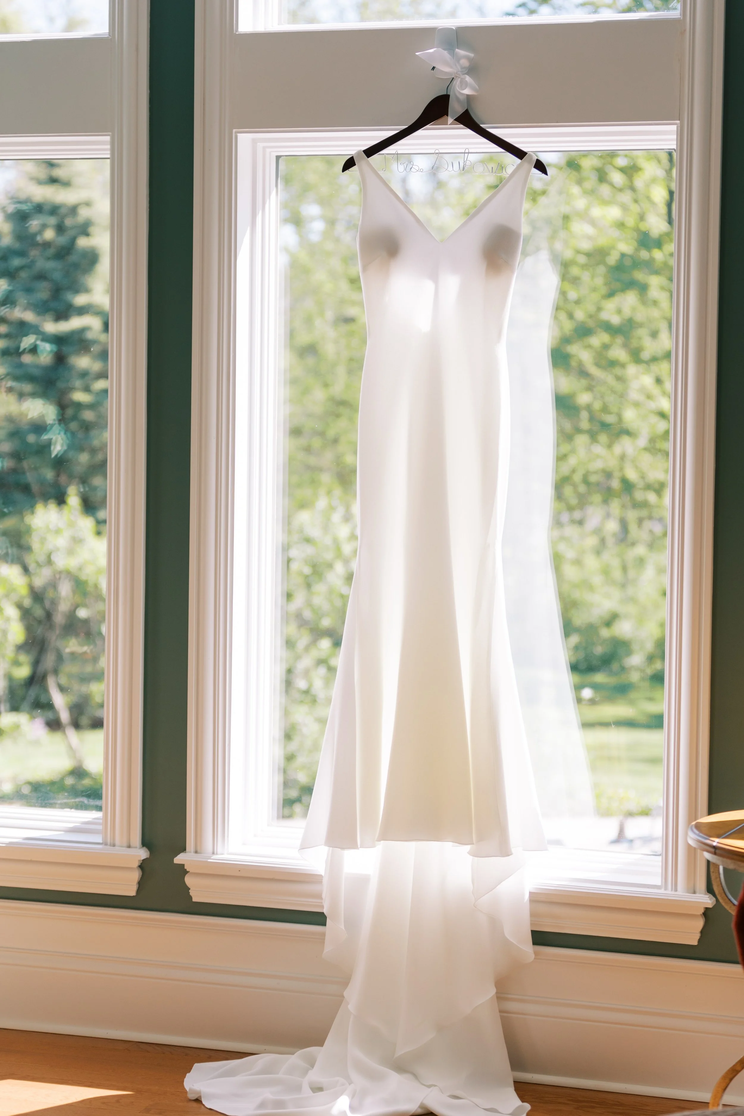 A white wedding dress hanging on a black hanger from a window with trees outside.