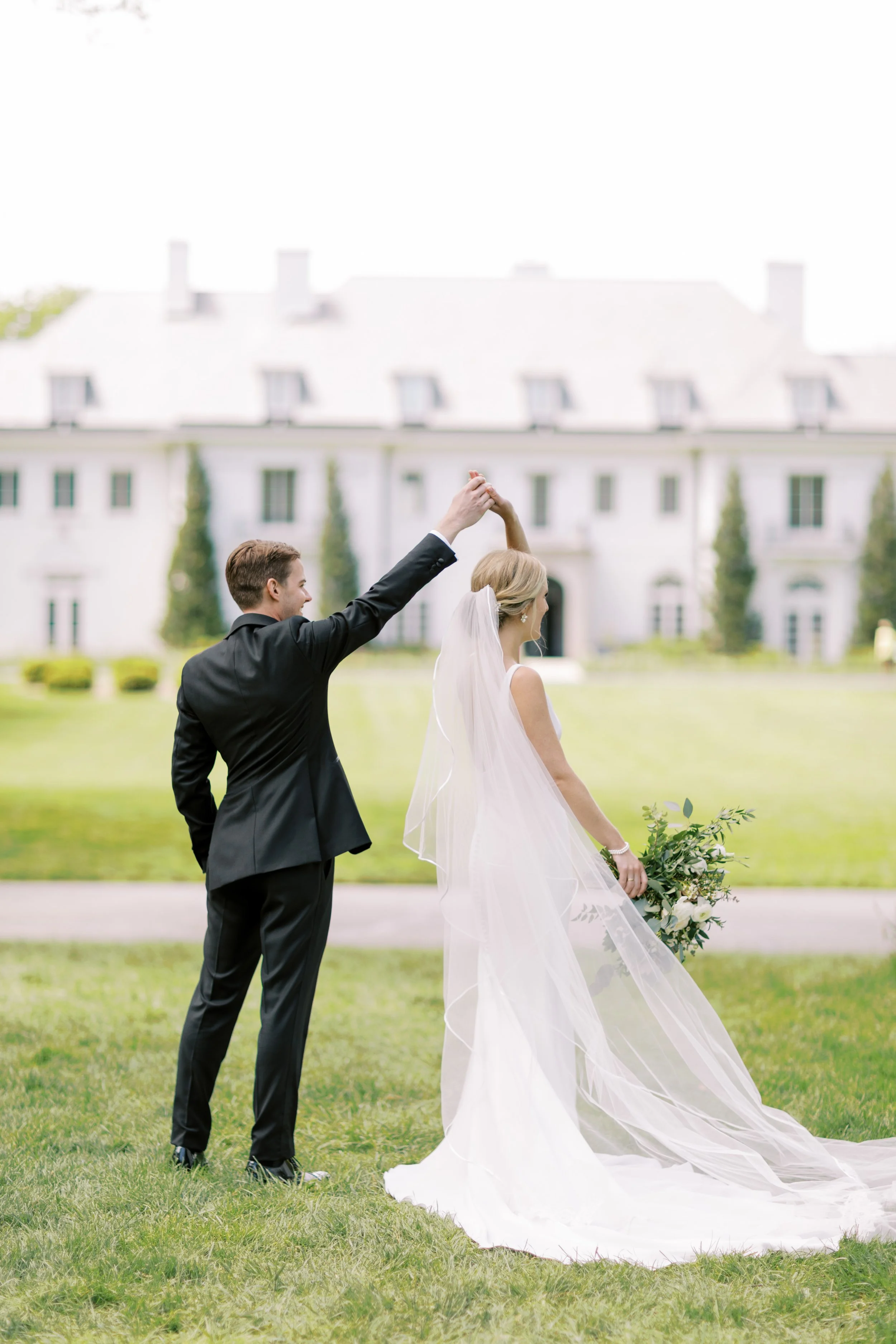 A newlywed couple dancing outside a large white house with green trees in the background. The groom is wearing a black tuxedo and the bride a white wedding dress with a veil, holding a bouquet of white flowers.