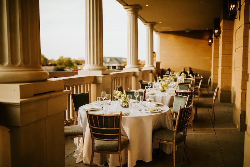 Elegantly set outdoor dining tables on a spacious balcony with white tablecloths, glassware, silverware, floral centerpieces, surrounded by chairs, with large columns and warm lighting, during sunset.