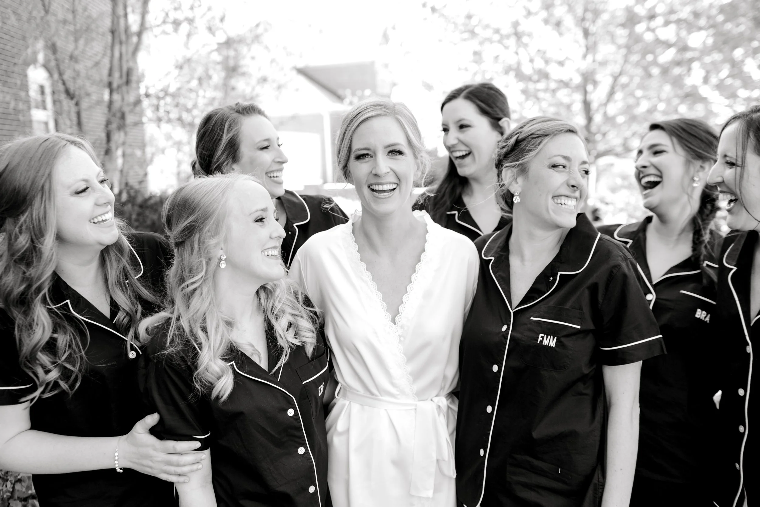 Group of women wearing matching pajamas, smiling, and laughing outdoors in a park setting, with trees and a building in the background.