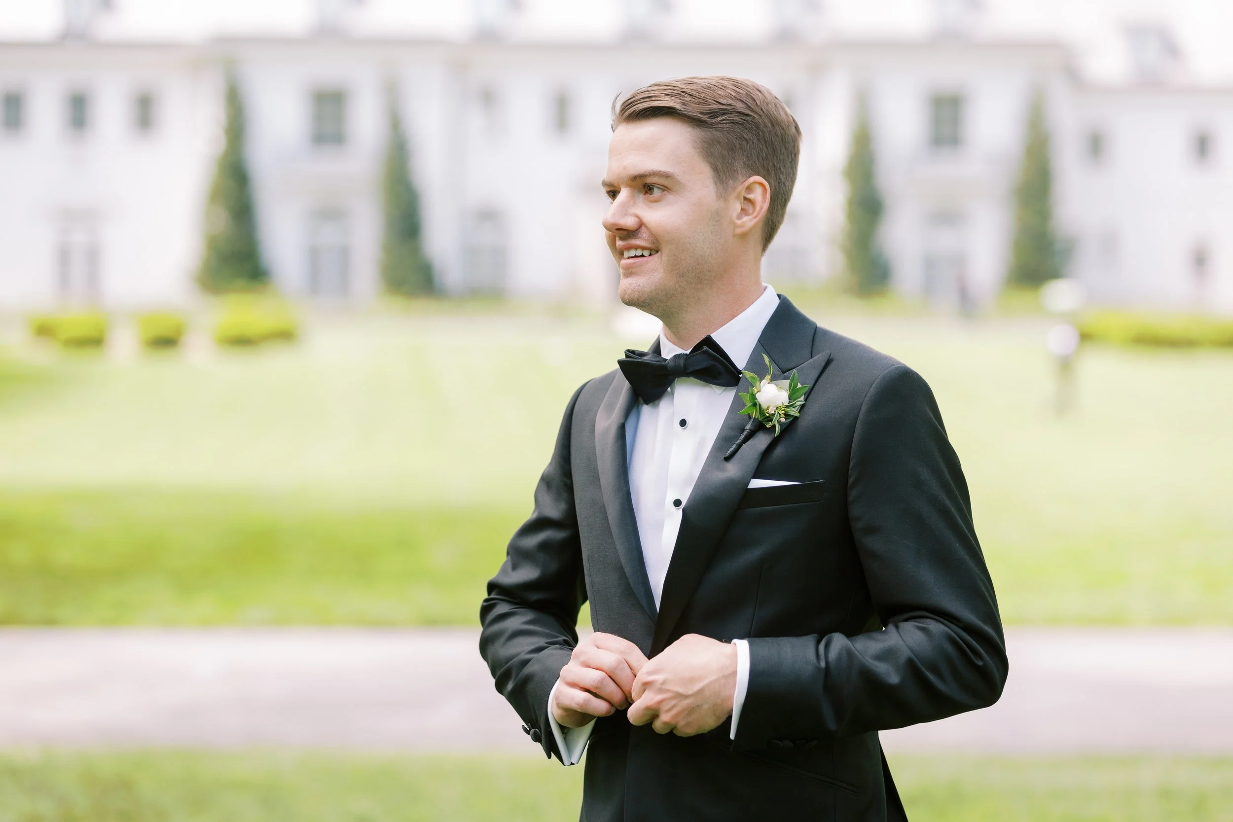 A man dressed in a black tuxedo with a white shirt and black bow tie, standing outdoors in front of a blurred background of a large building and greenery. He has short, styled brown hair and is smiling slightly, holding his hands together in front of