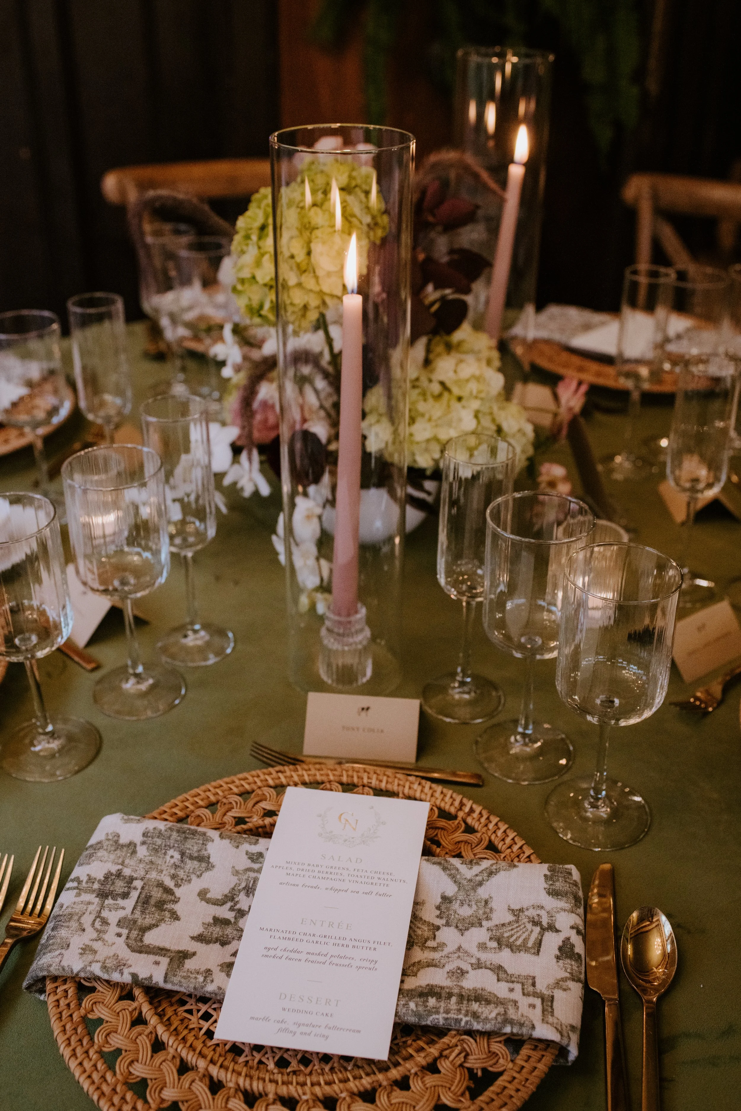 A decorated dining table with wine glasses, candles, floral centerpieces, and place settings. Included is a menu card on a napkin-covered basket at each place setting.