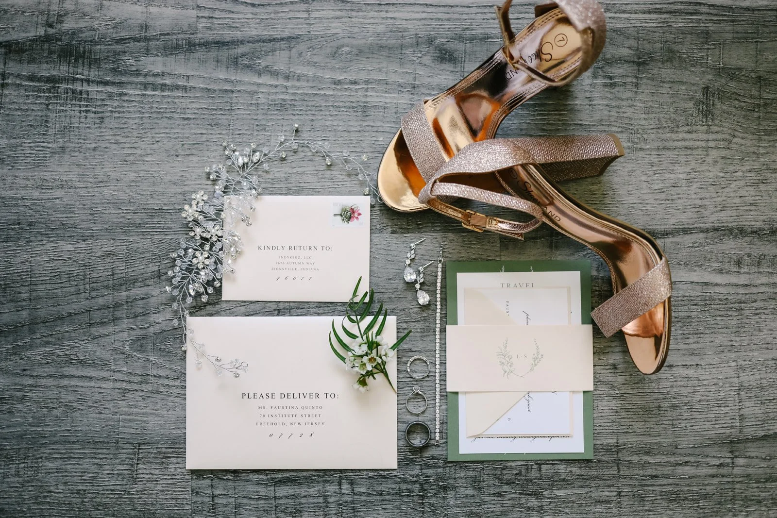 Wedding accessories on a rustic wooden surface, including a pair of rose gold high-heeled shoes, a bouquet of white flowers, a pair of earrings, a bracelet, a ring, a jewelry necklace, and wedding invitation cards.