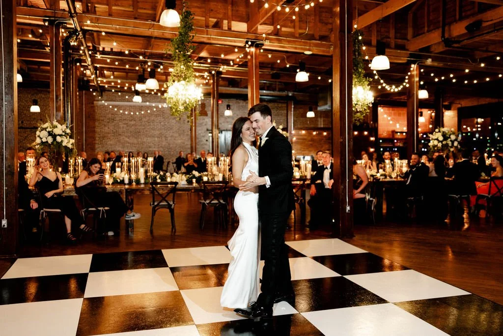 A newlywed couple sharing their first dance at a wedding reception in a rustic indoor venue with string lights and floral decorations