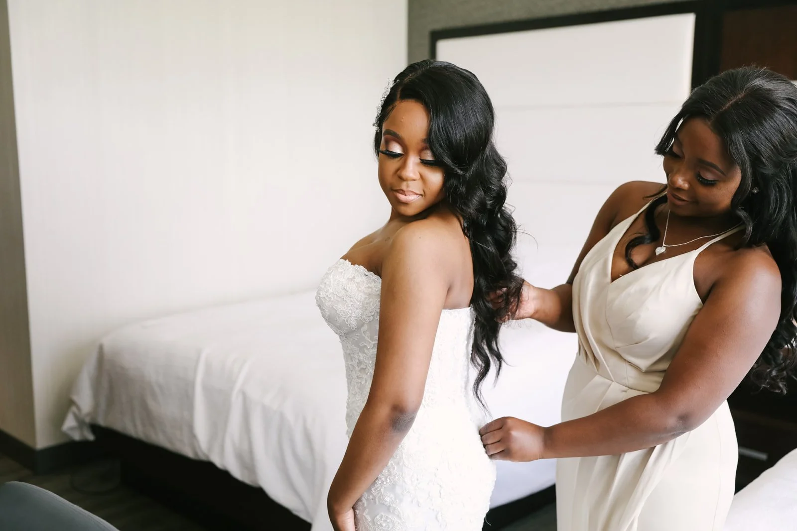 A bride in a strapless white wedding dress is having her dress adjusted by a woman in a cream-colored dress, in a bedroom with a bed and a framed picture on the wall.