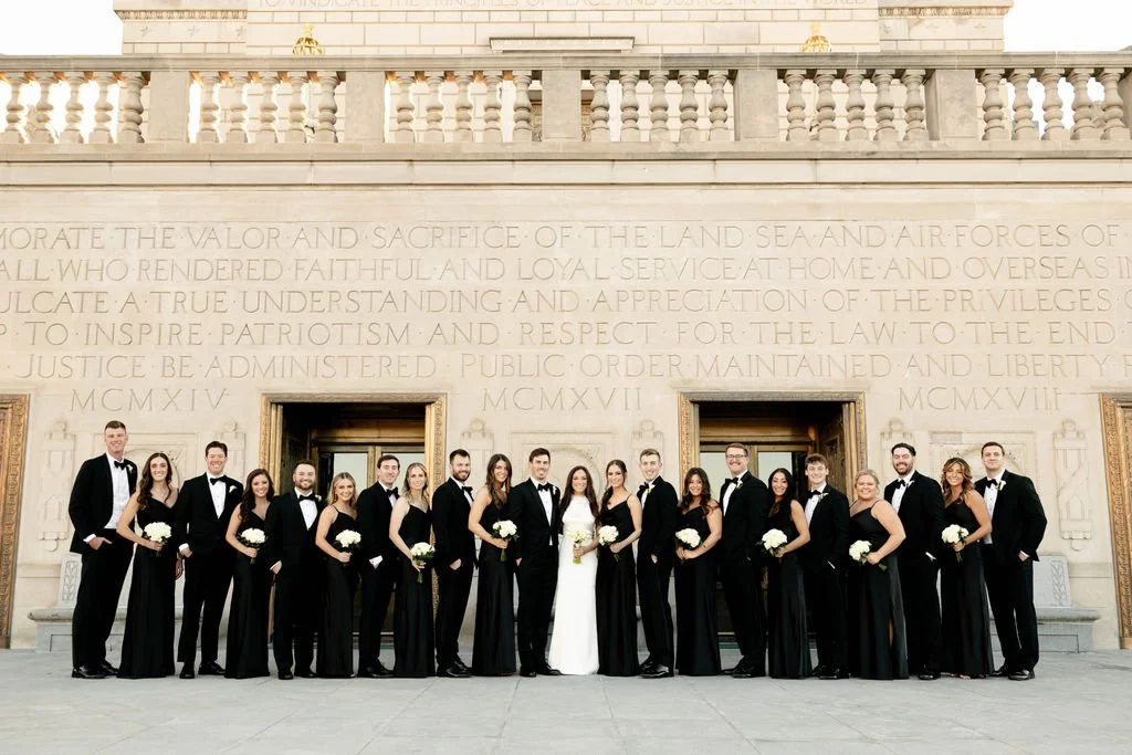 A wedding party posing in front of a monument with engraved text, featuring a bride in a white gown and bridesmaids in black dresses, and groomsmen in black suits with bow ties.